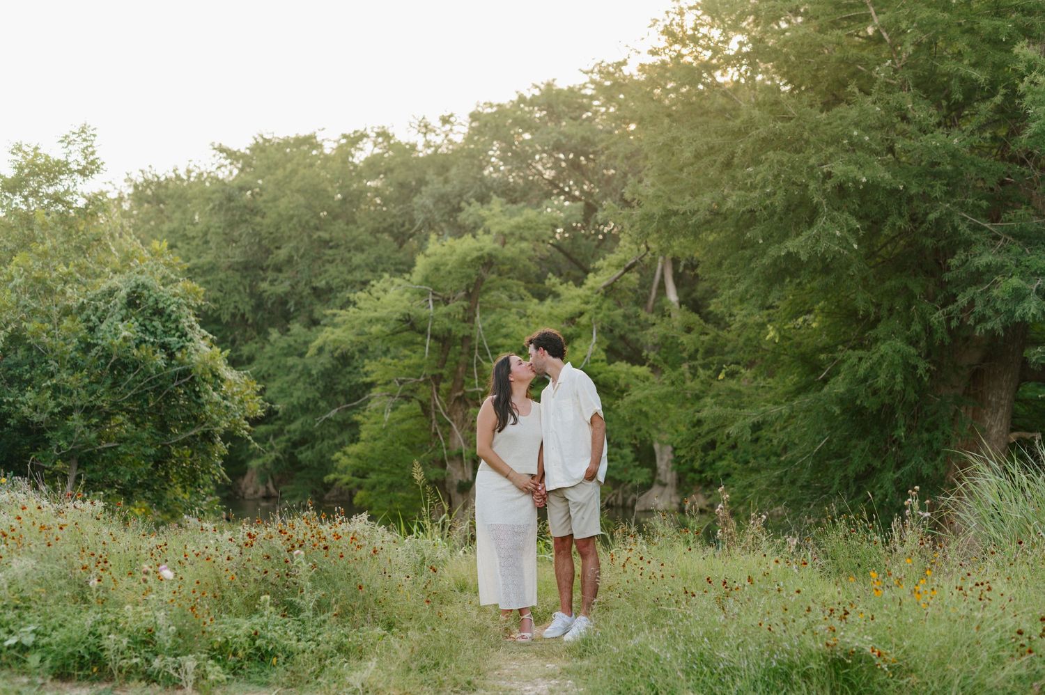 Couple embraces playfully in a lush green outdoor setting during golden hour.
