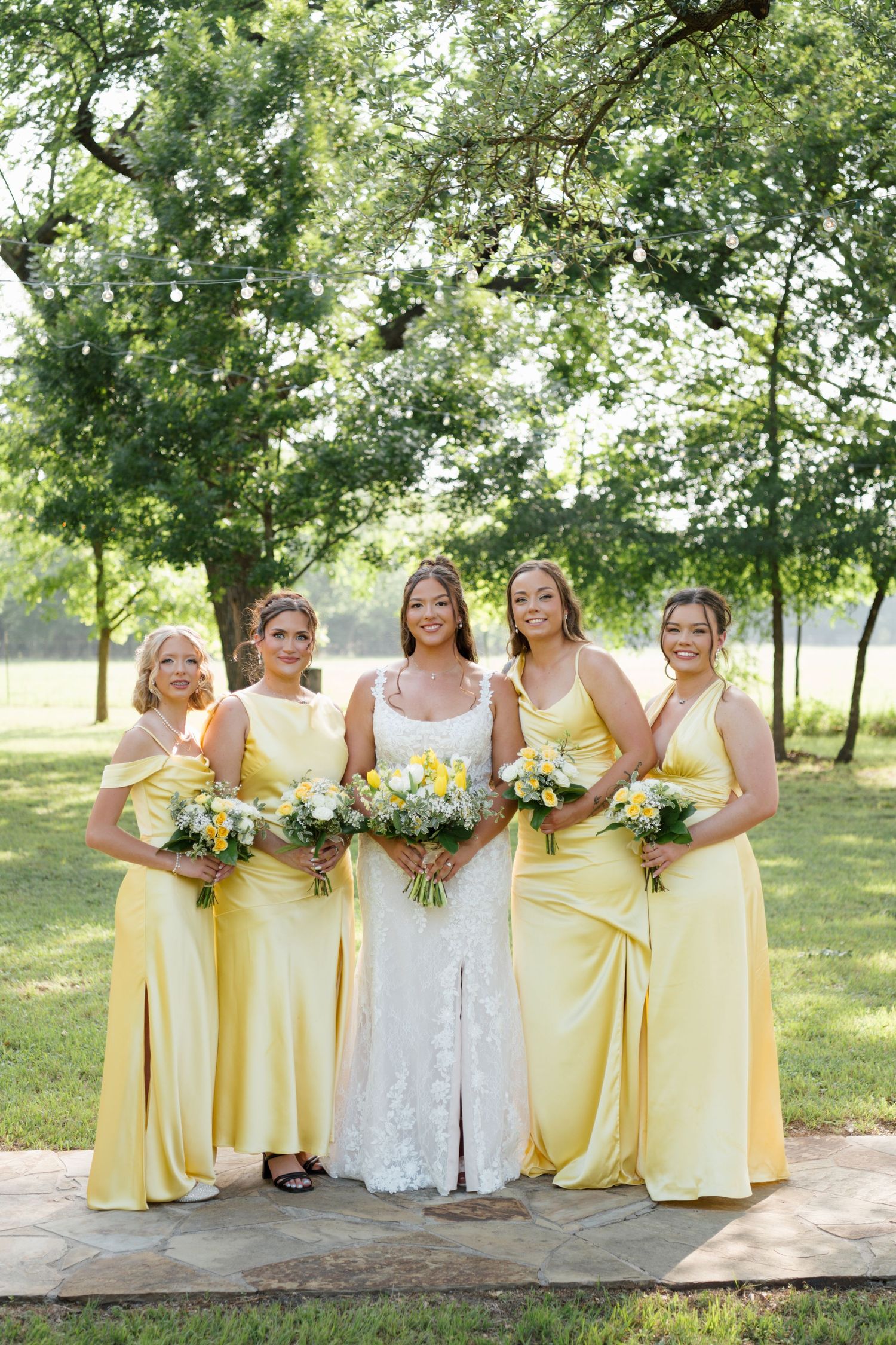 Bride in lace gown poses with four bridesmaids in yellow dresses holding bouquets outdoors.