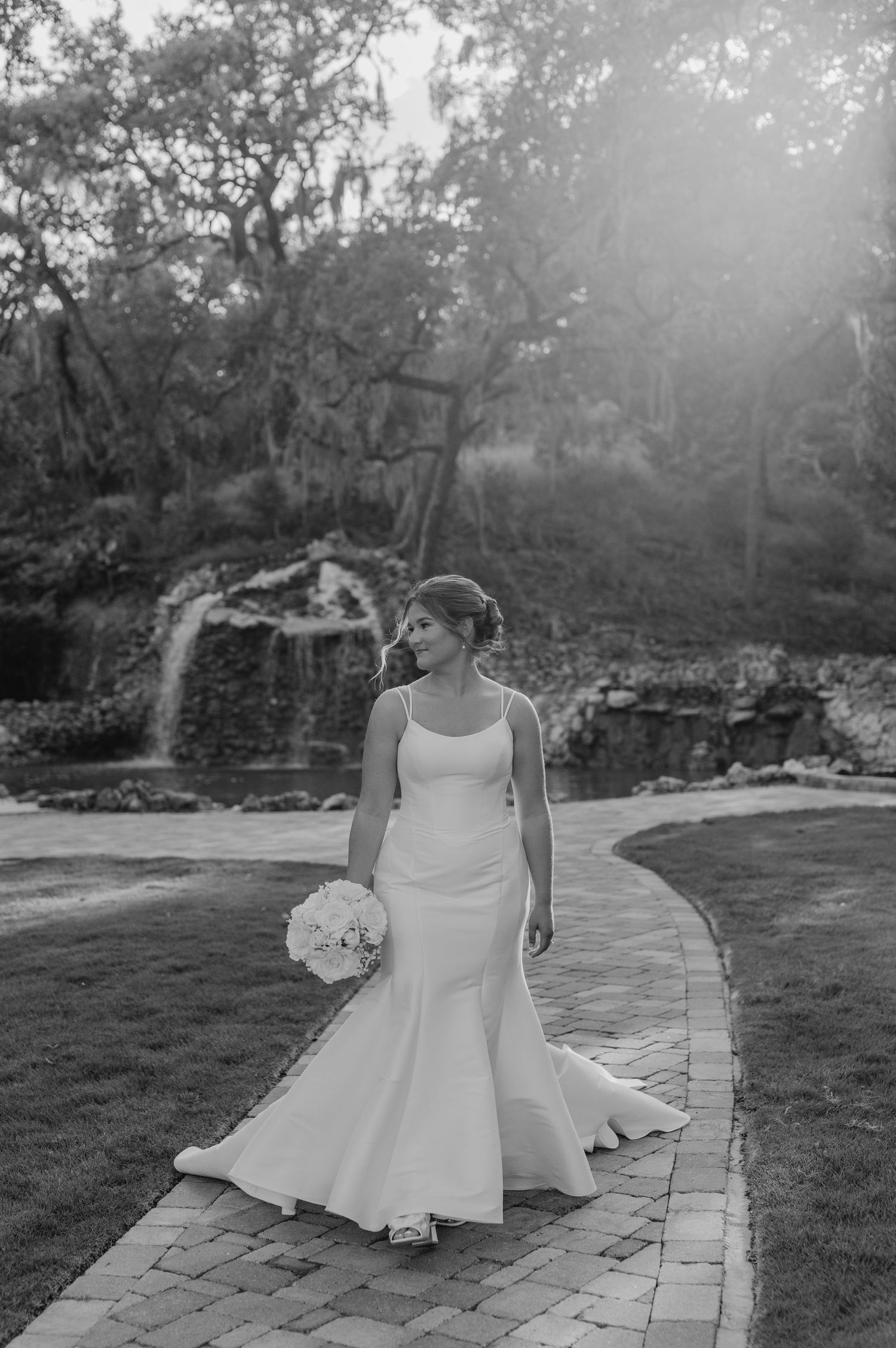 A classic white wedding dress photographed in black and white on a stone pathway in a garden setting.