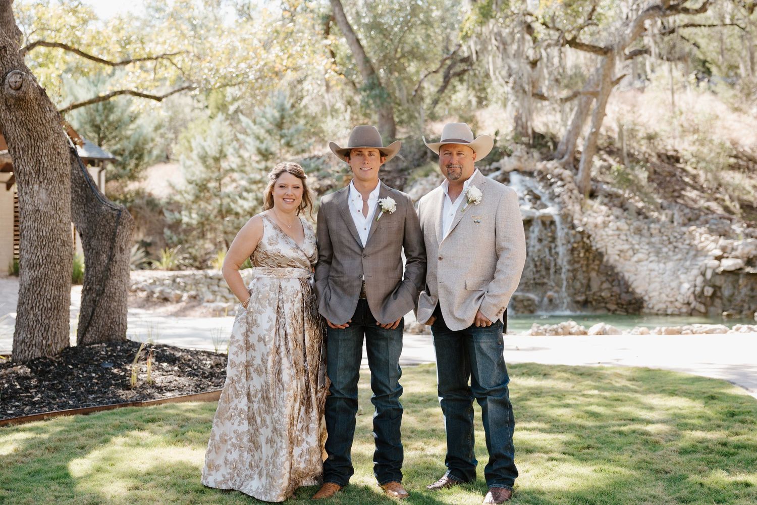Wedding party in cowboy attire poses outdoors under trees at a rustic venue.