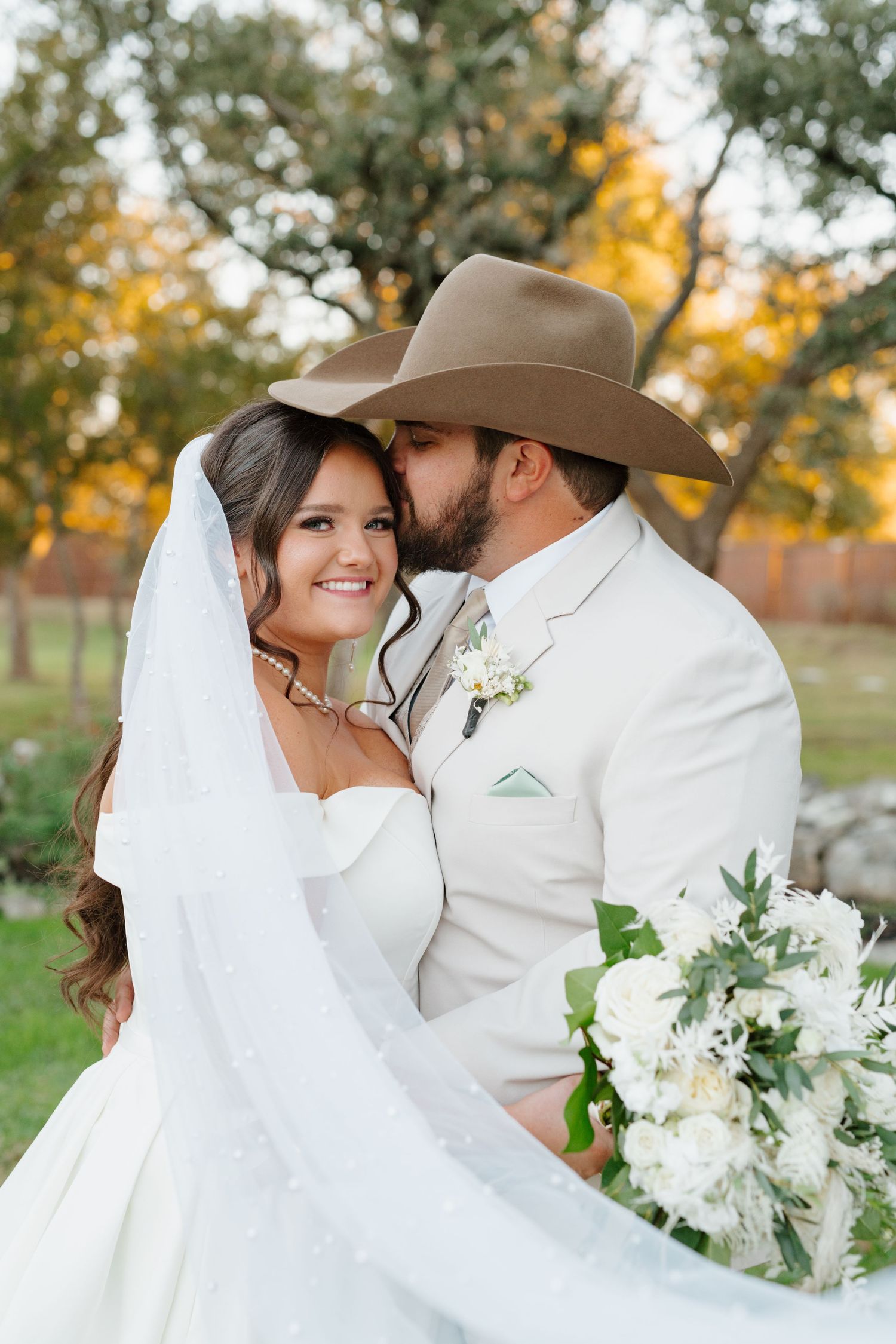 A romantic country wedding couple embraces outdoors with white bouquet and cowboy hat against autumn foliage.