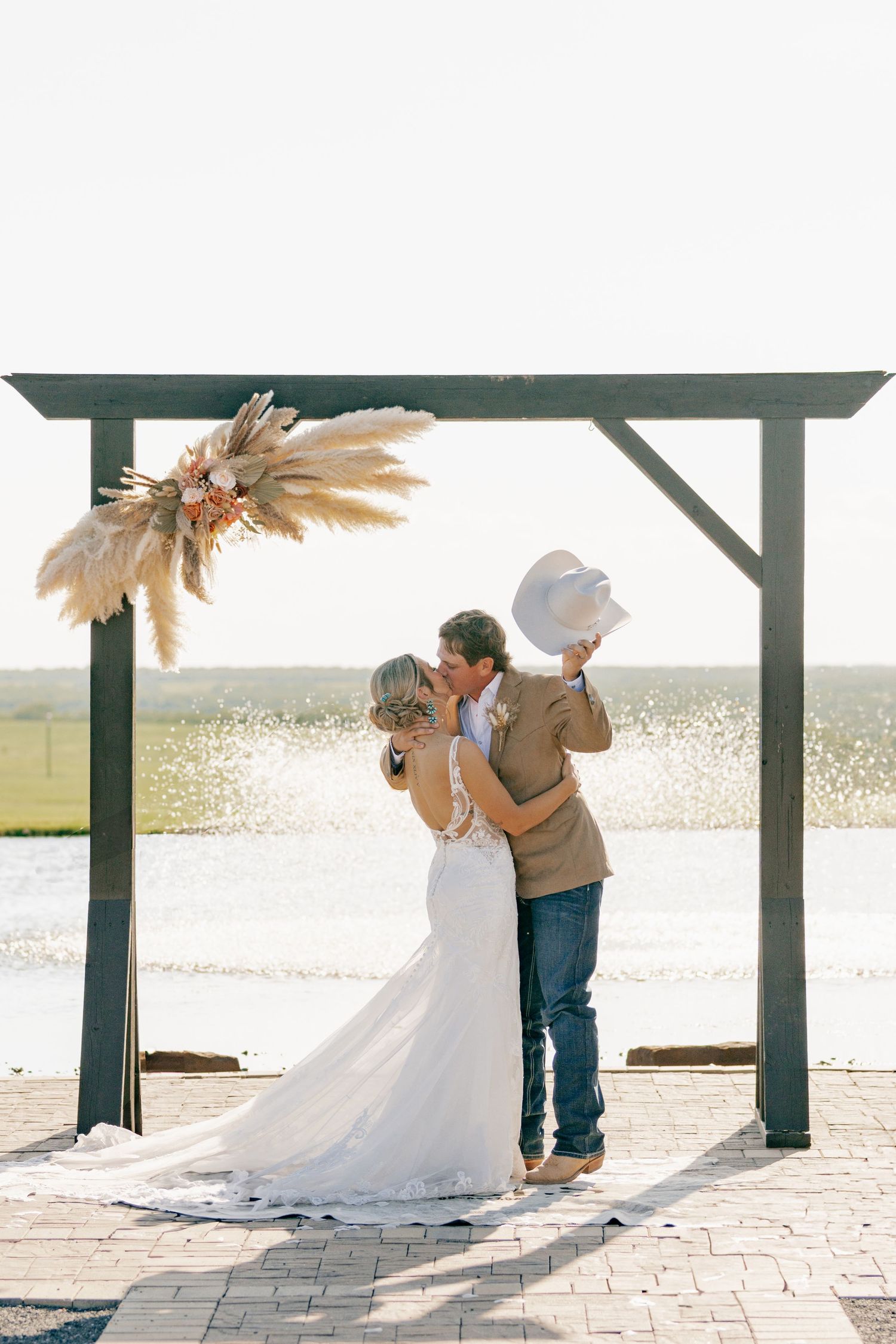 A couple shares their first kiss under a wooden arch decorated with pampas grass at sunset.