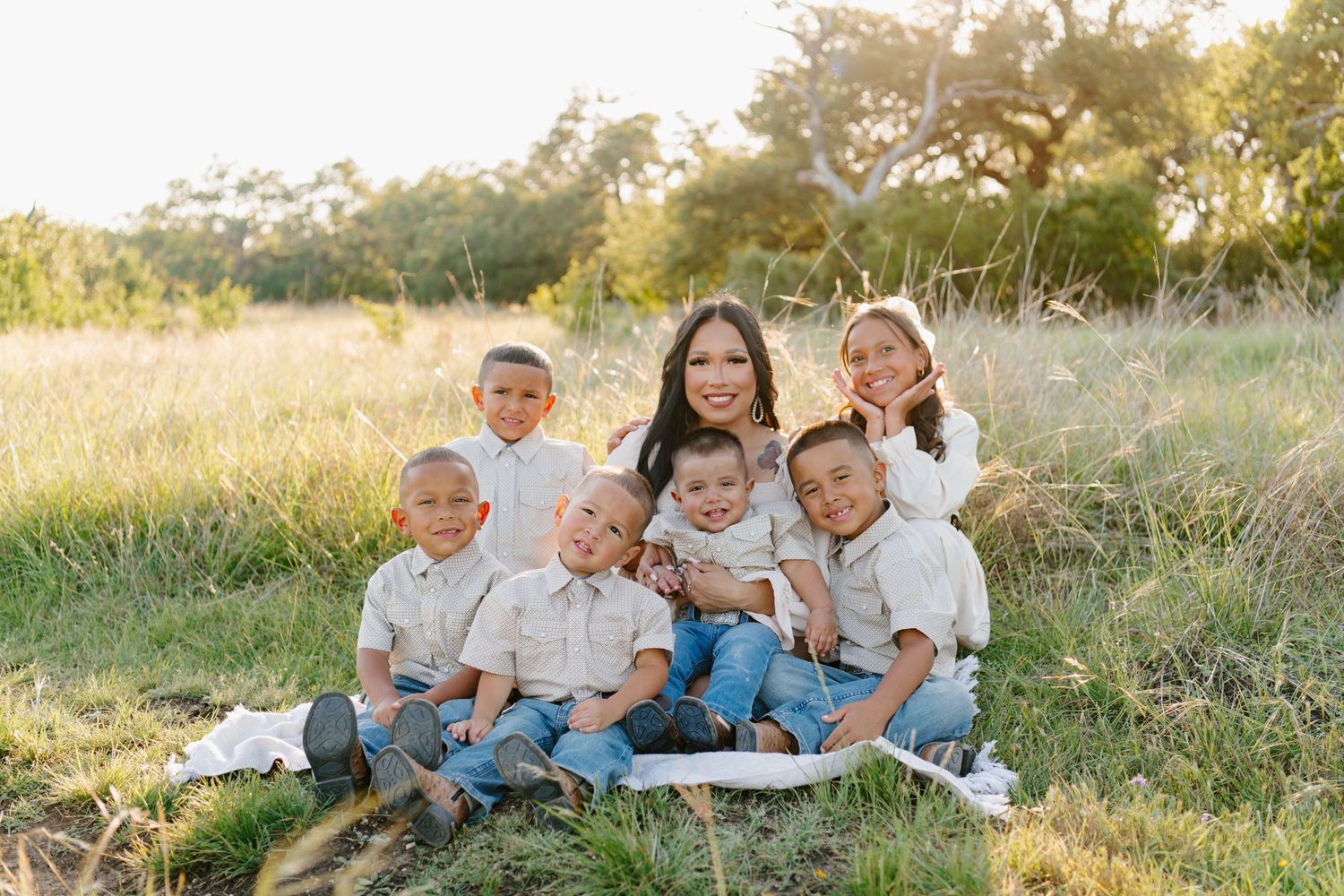 Family group sitting together on blanket in sun-drenched field with tall grass and trees during golden hour.