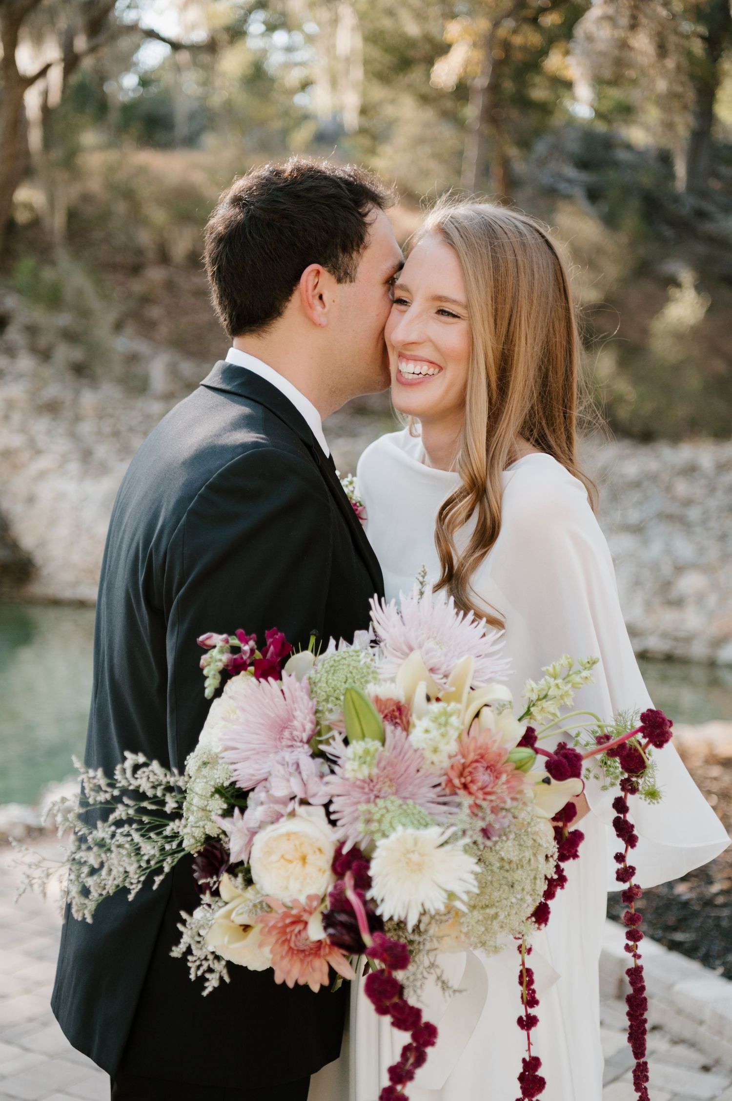 Wedding bouquet with burgundy and pink flowers against an outdoor natural backdrop.