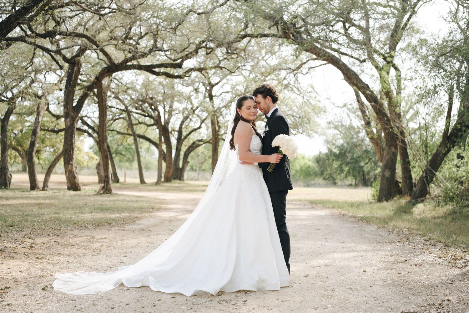 Bride and groom embracing on a scenic tree-lined path in wedding attire for romantic outdoor portraits.