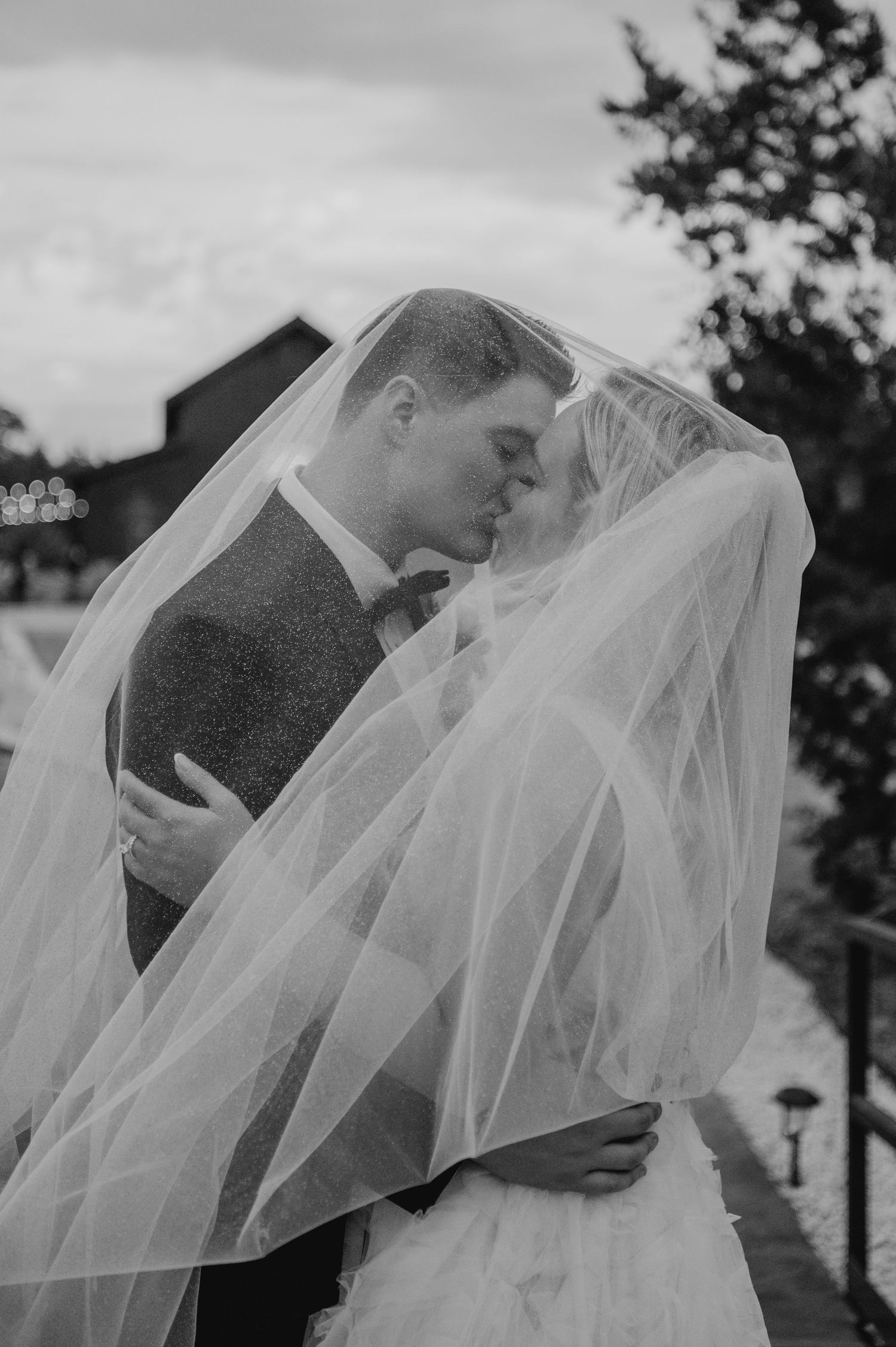 A black and white photograph of a bride and groom embracing each other in a romantic outdoor wedding setting.