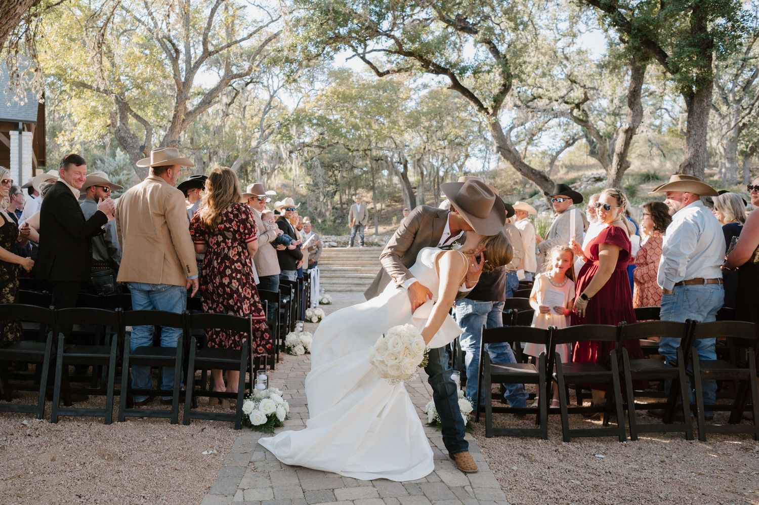 Wedding ceremony guests seated outdoors under large oak trees during a sunny day.