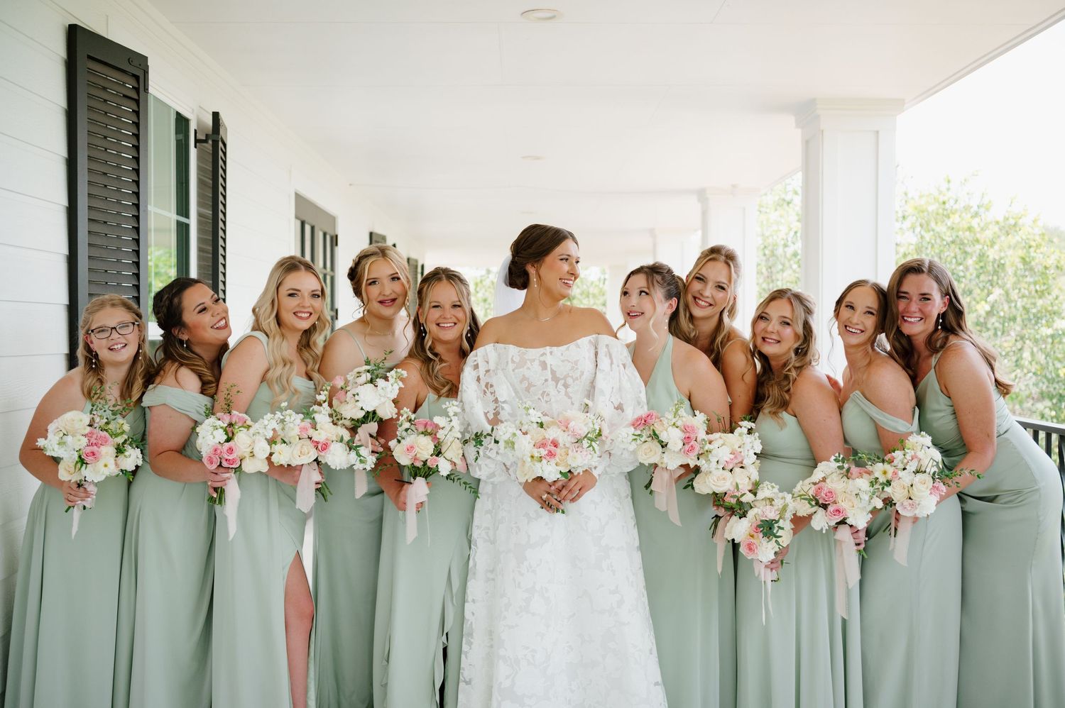 Row of bridesmaids in sage green dresses holding pink and white bouquets on a bright white porch.