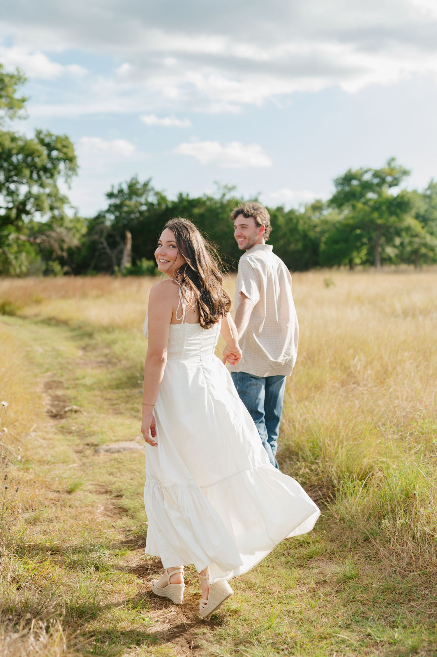 A couple strolls hand in hand along a winding dirt path through golden grass fields.