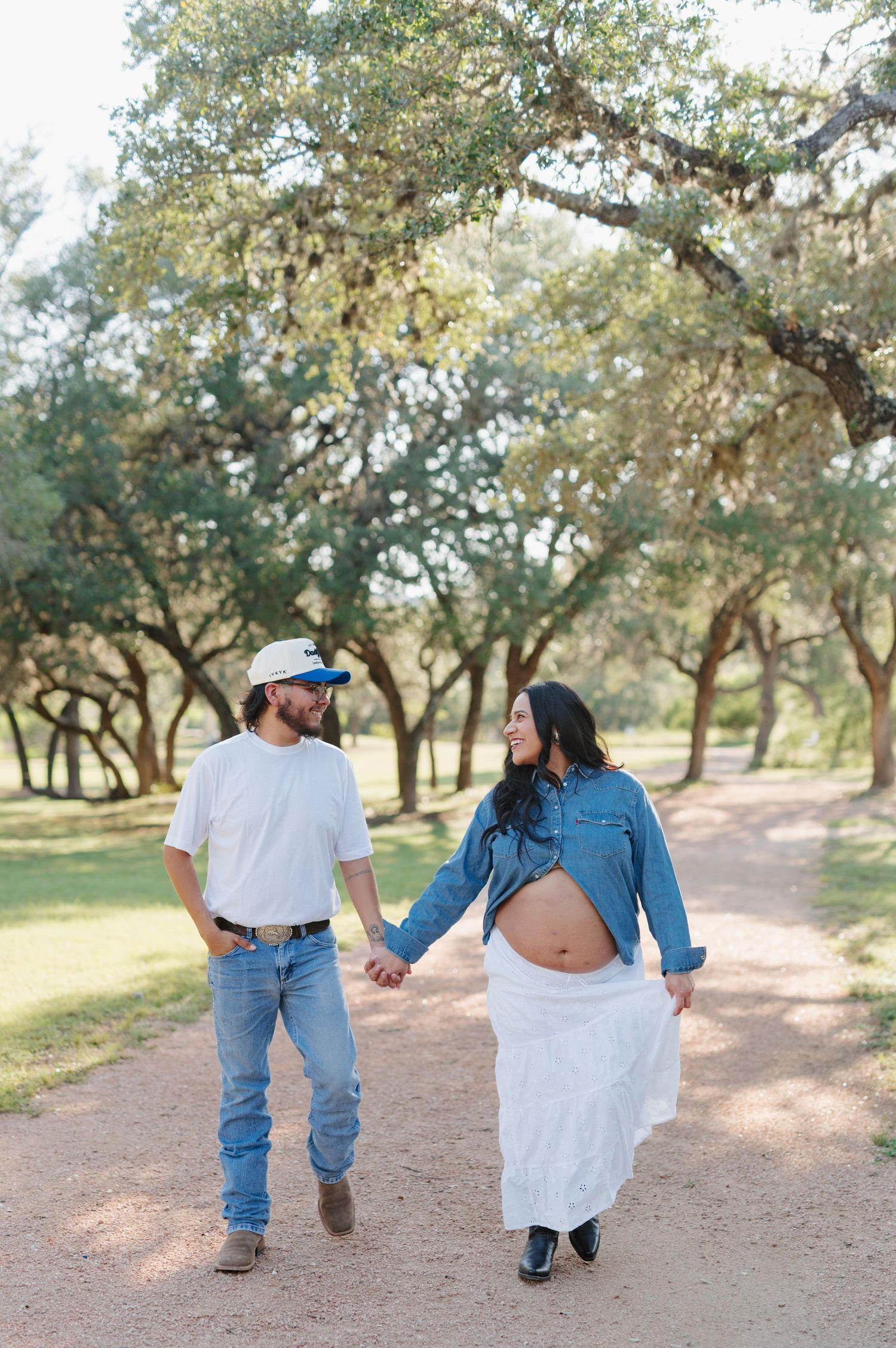 A romantic couple walks hand in hand through an olive grove on a sunny afternoon wearing denim and white.