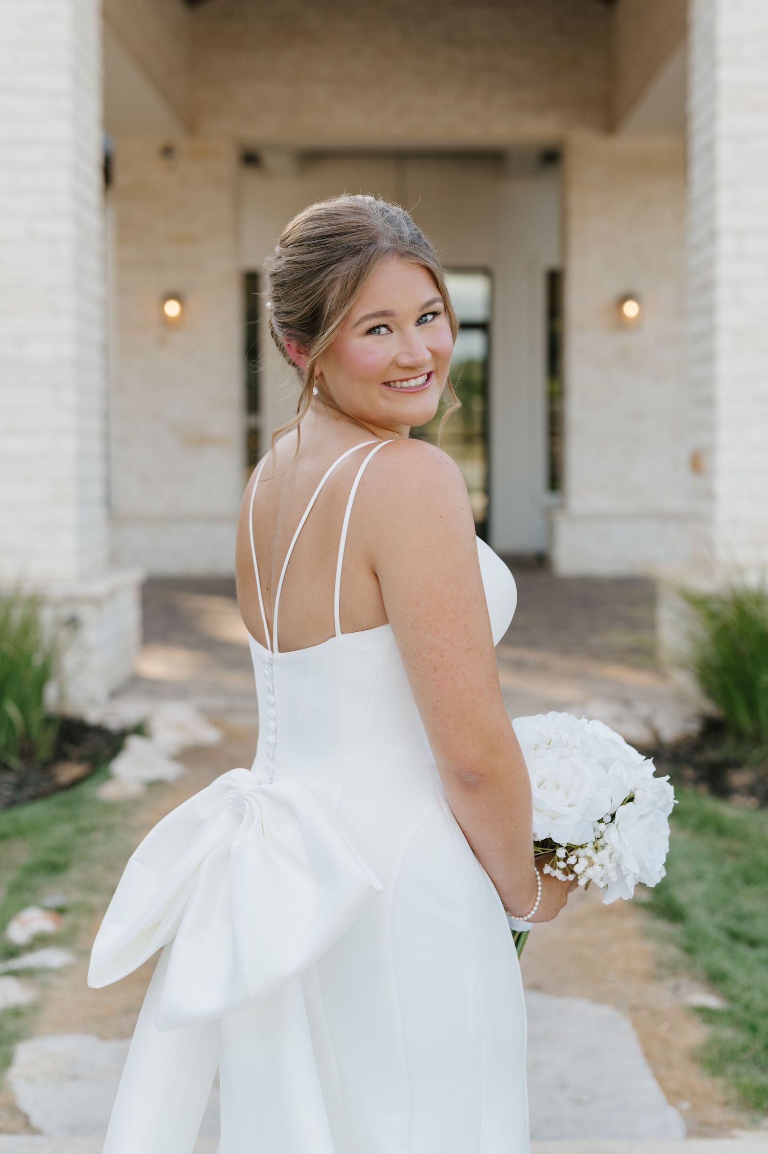 Bride in a sleek white dress with straps poses against a light stone building backdrop.