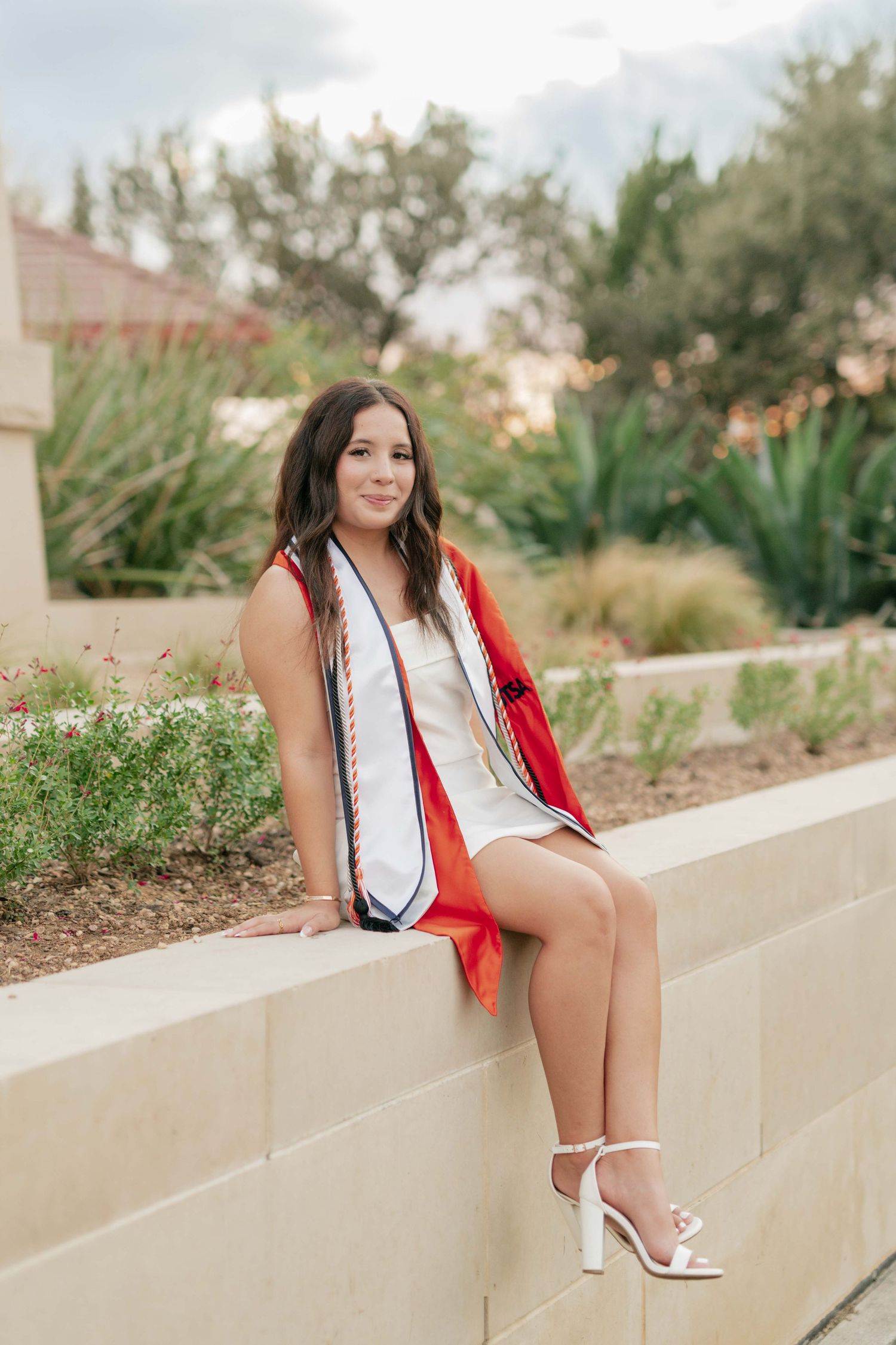 Graduate in graduation attire sits on concrete wall with desert garden backdrop.