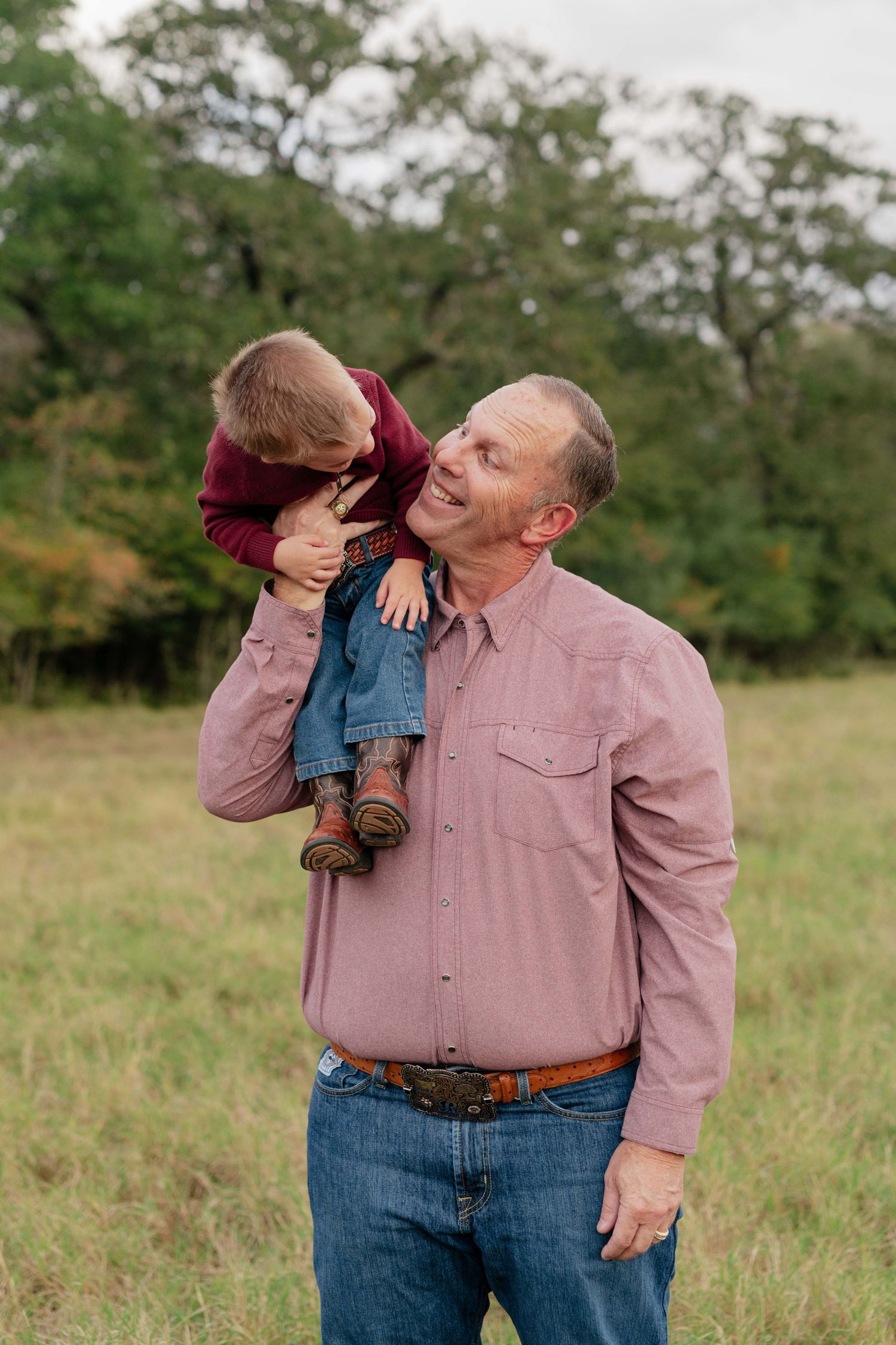 Adult and small child share playful moment in casual outdoor setting with grass and trees.