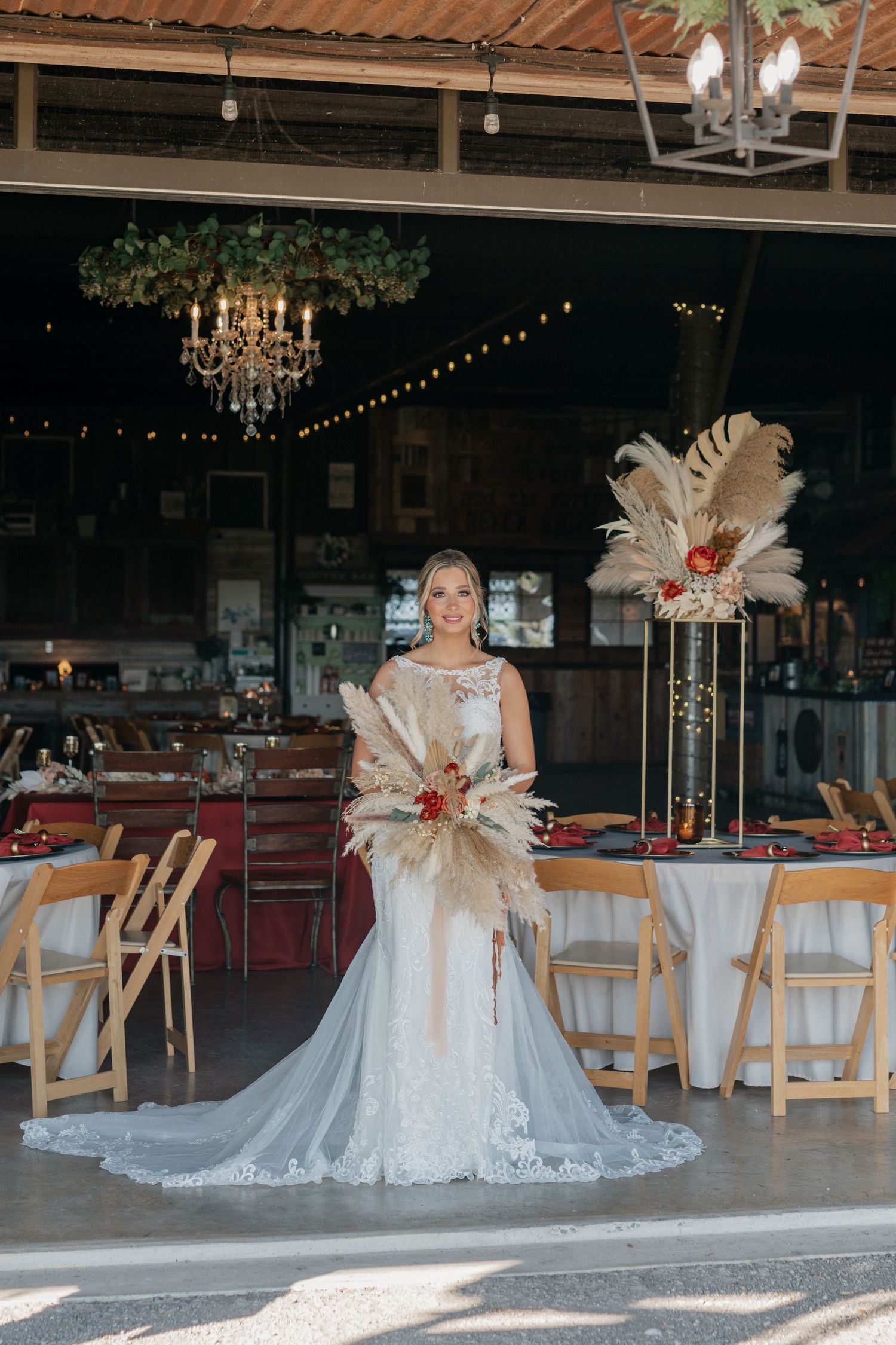 Bride in lace wedding dress with feather bouquet standing in rustic reception venue.