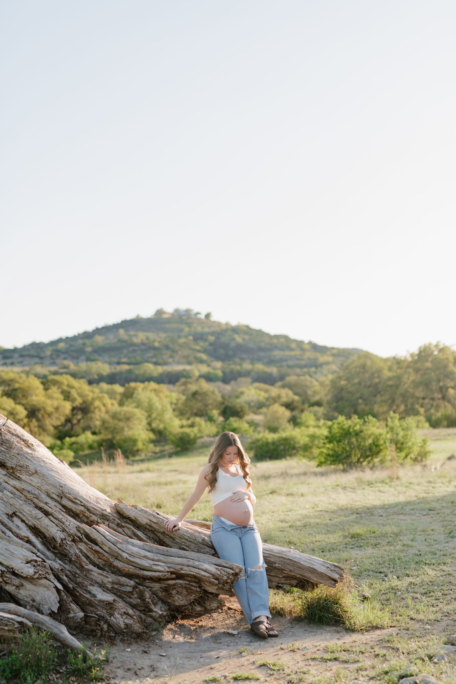 A person sits on a large fallen tree trunk against a backdrop of rolling green hills at sunset.