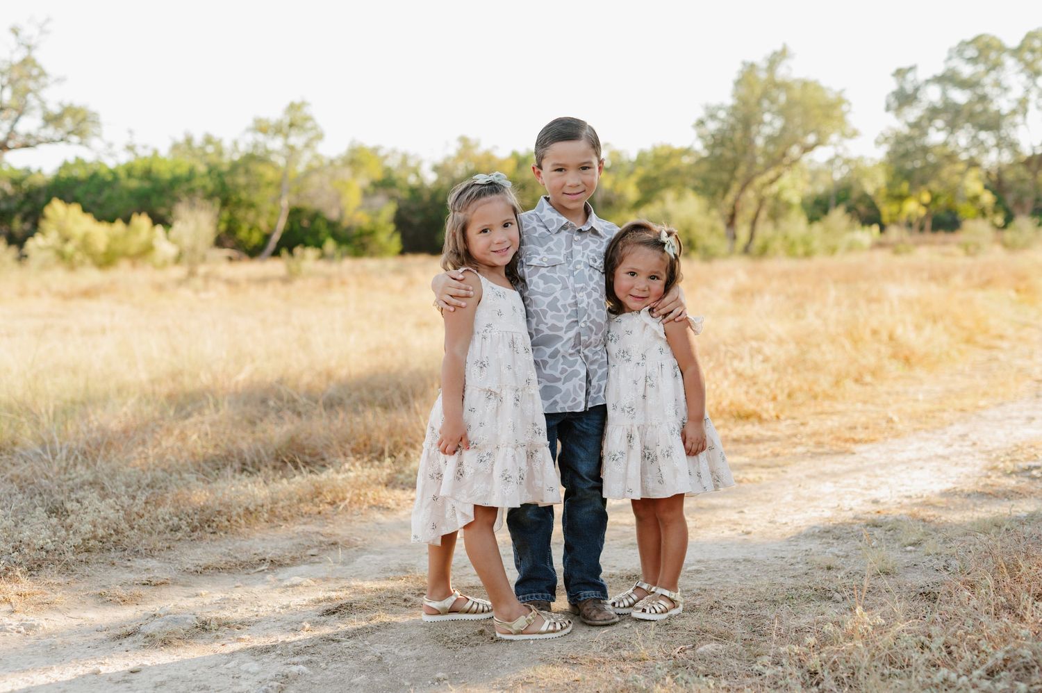 A family poses together on a sunny dirt path surrounded by golden grass and trees in a natural outdoor setting.