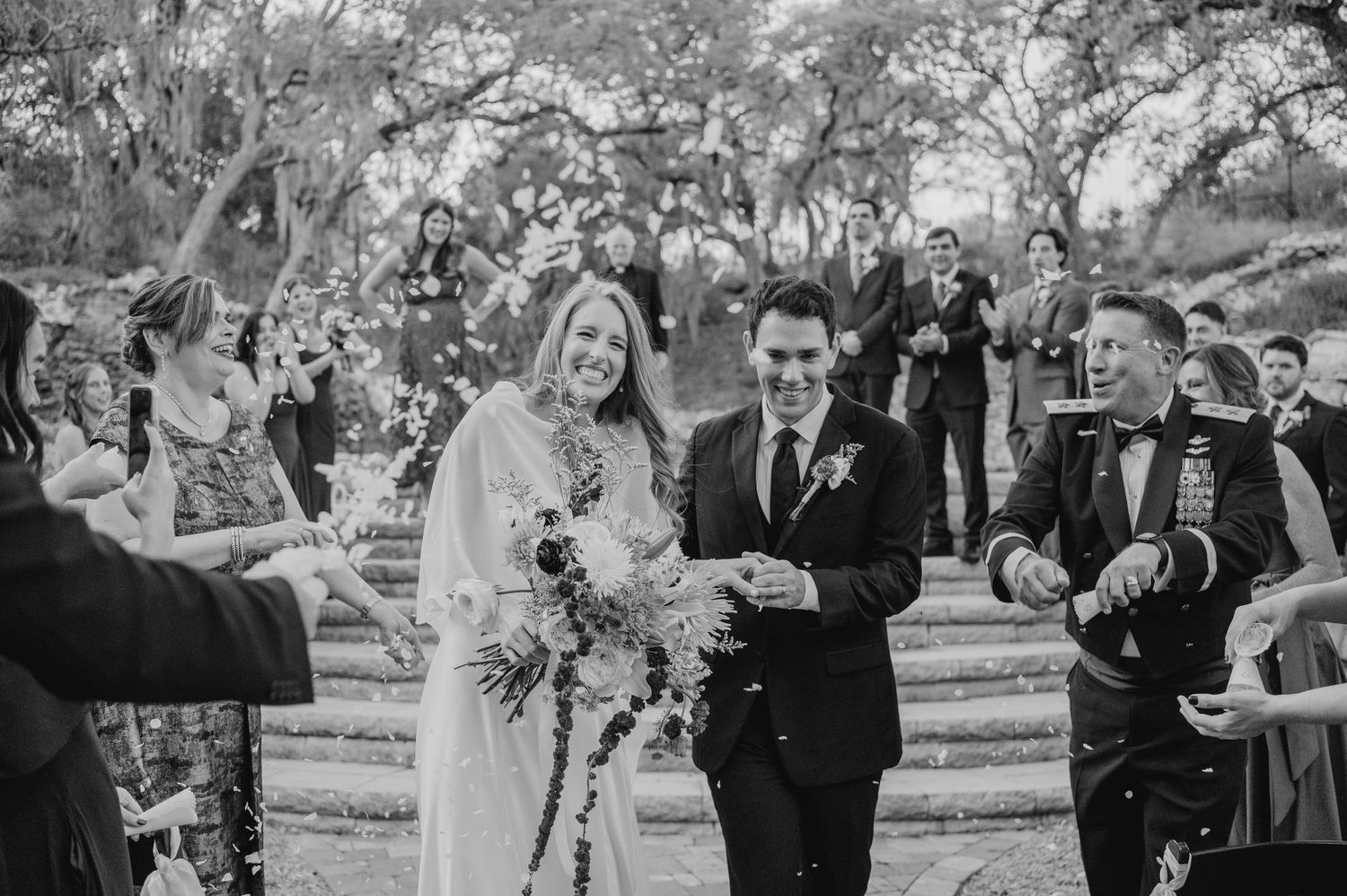 Wedding celebration with guests tossing confetti at a joyful outdoor ceremony under trees in black and white.