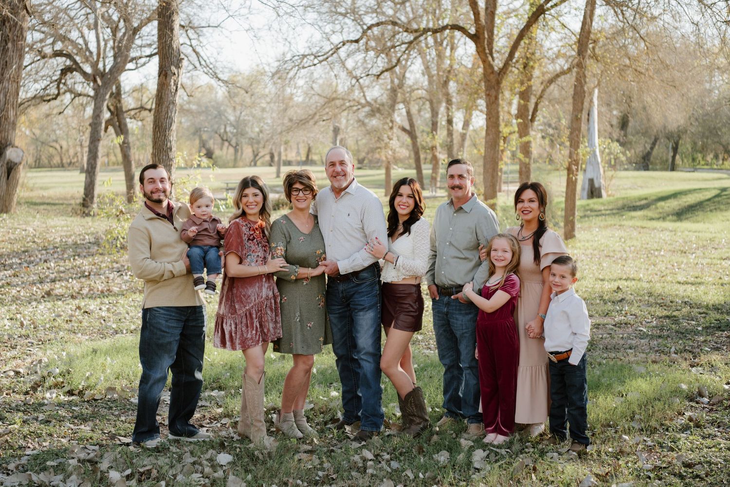 Multi-generational family group standing together outdoors in a park with bare trees and scattered leaves on the ground.