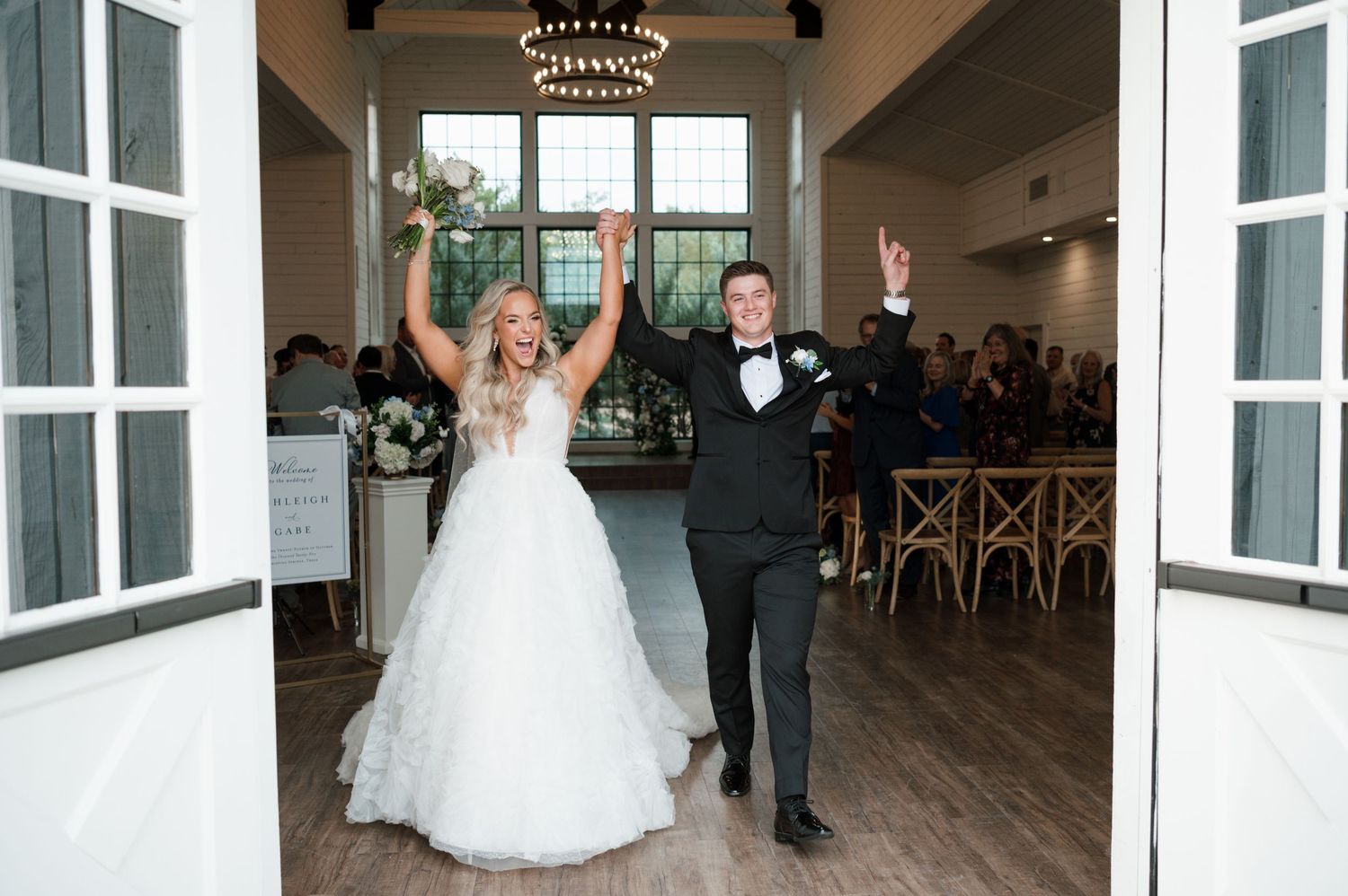 A bride and groom stand together in a formal wedding setting with large windows and a chandelier.