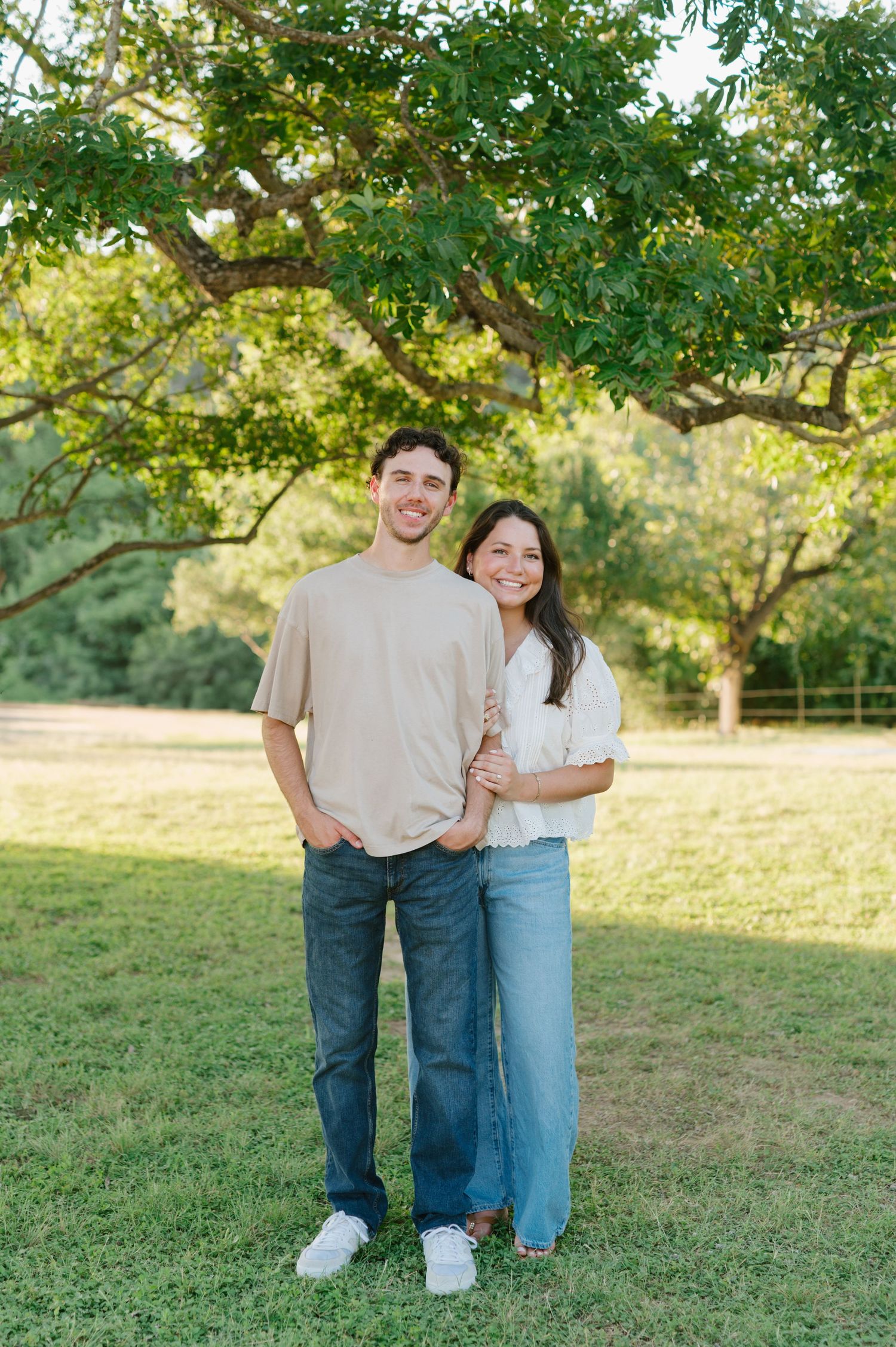 A casual couple in jeans posing together on green grass under trees.