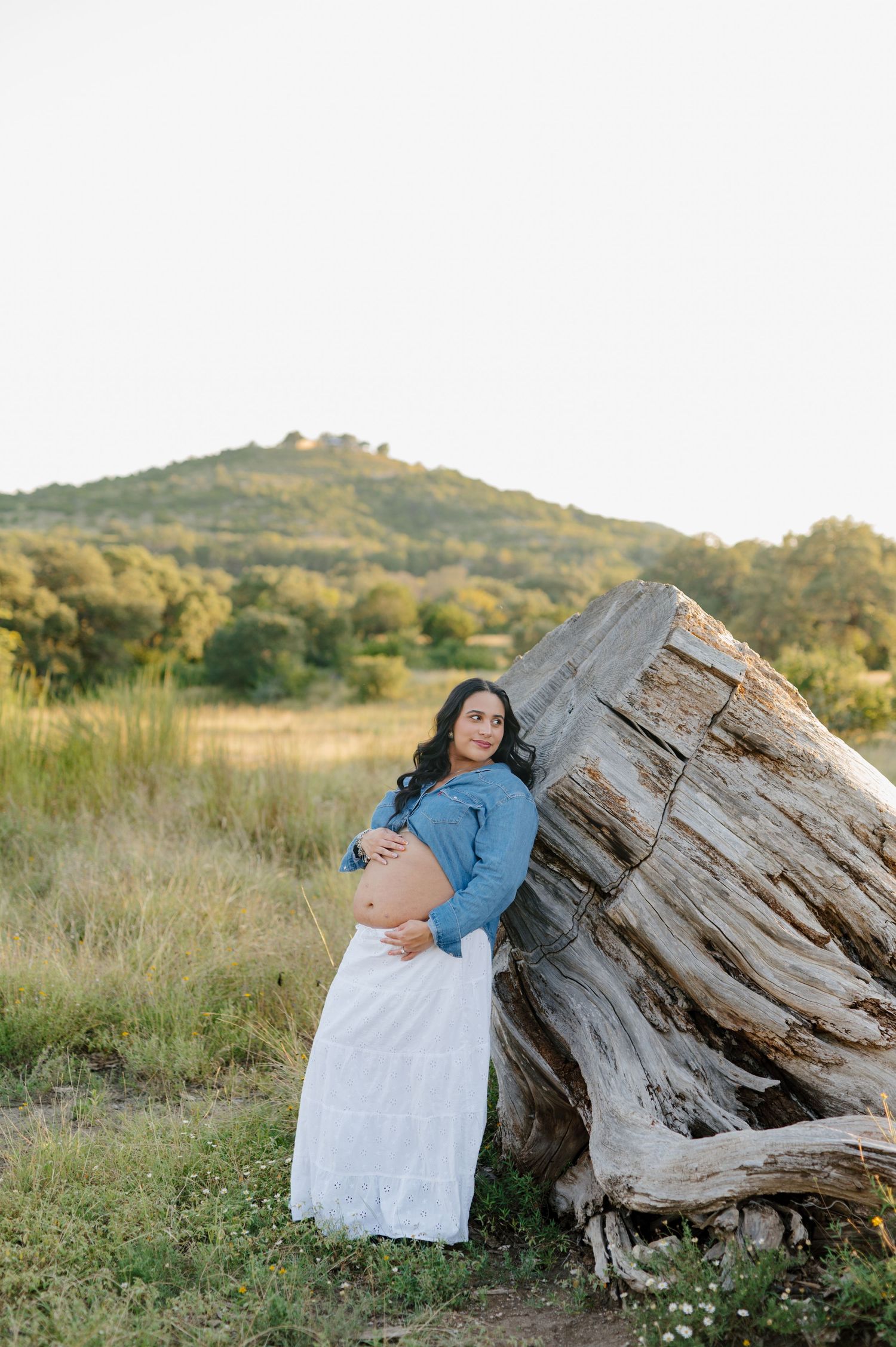 A woman in a white dress poses against a large weathered log in a grassy field at golden hour.
