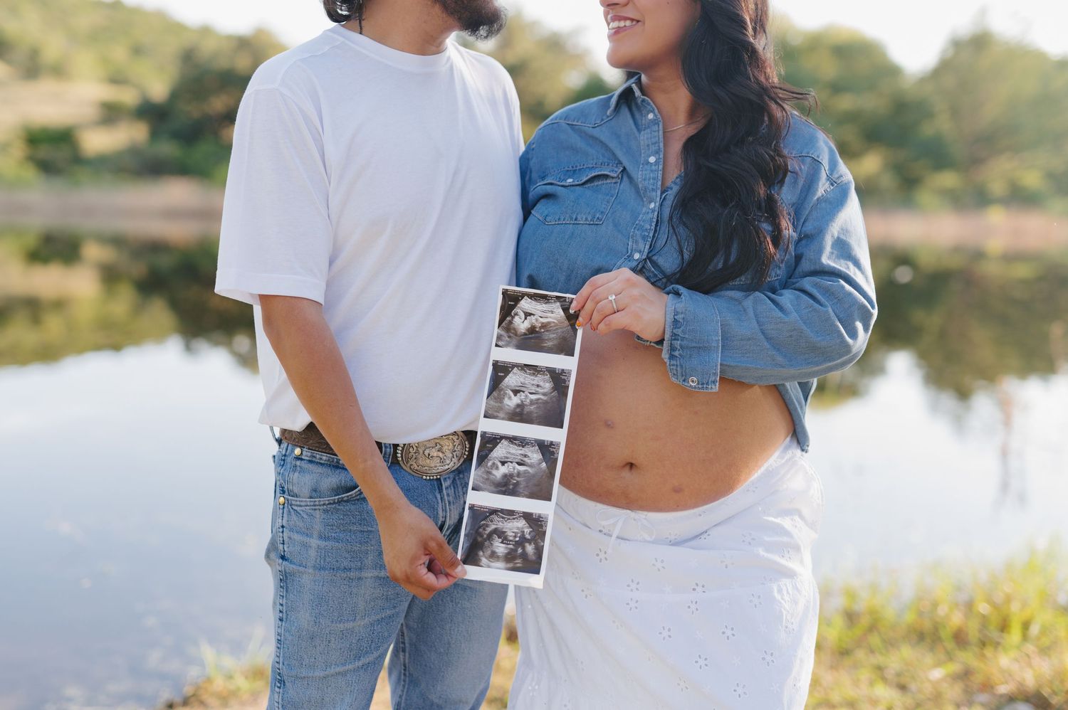 Couple holding ultrasound photo while embracing by scenic lakeside during sunset pregnancy announcement photoshoot.