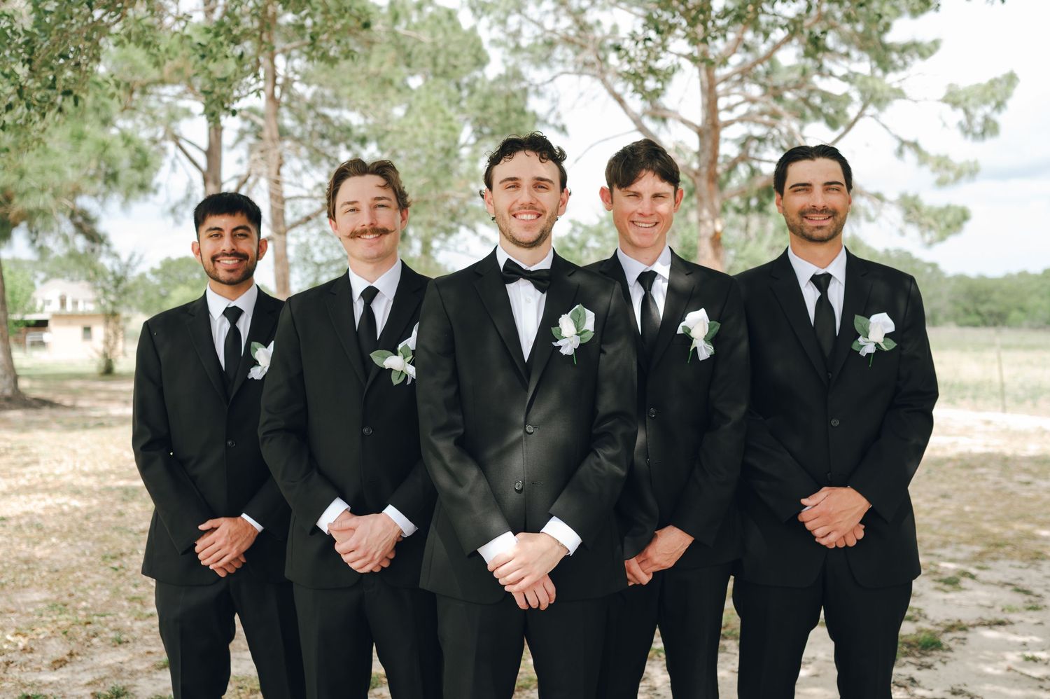 Five groomsmen in matching black tuxedos with white boutonnieres pose outdoors at a wedding ceremony.