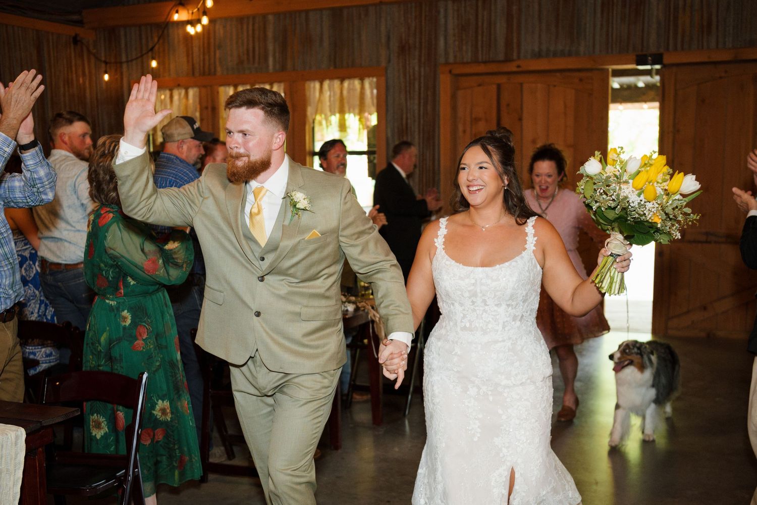 Joyful bride and groom walk hand-in-hand into rustic barn reception as guests cheer and applaud.