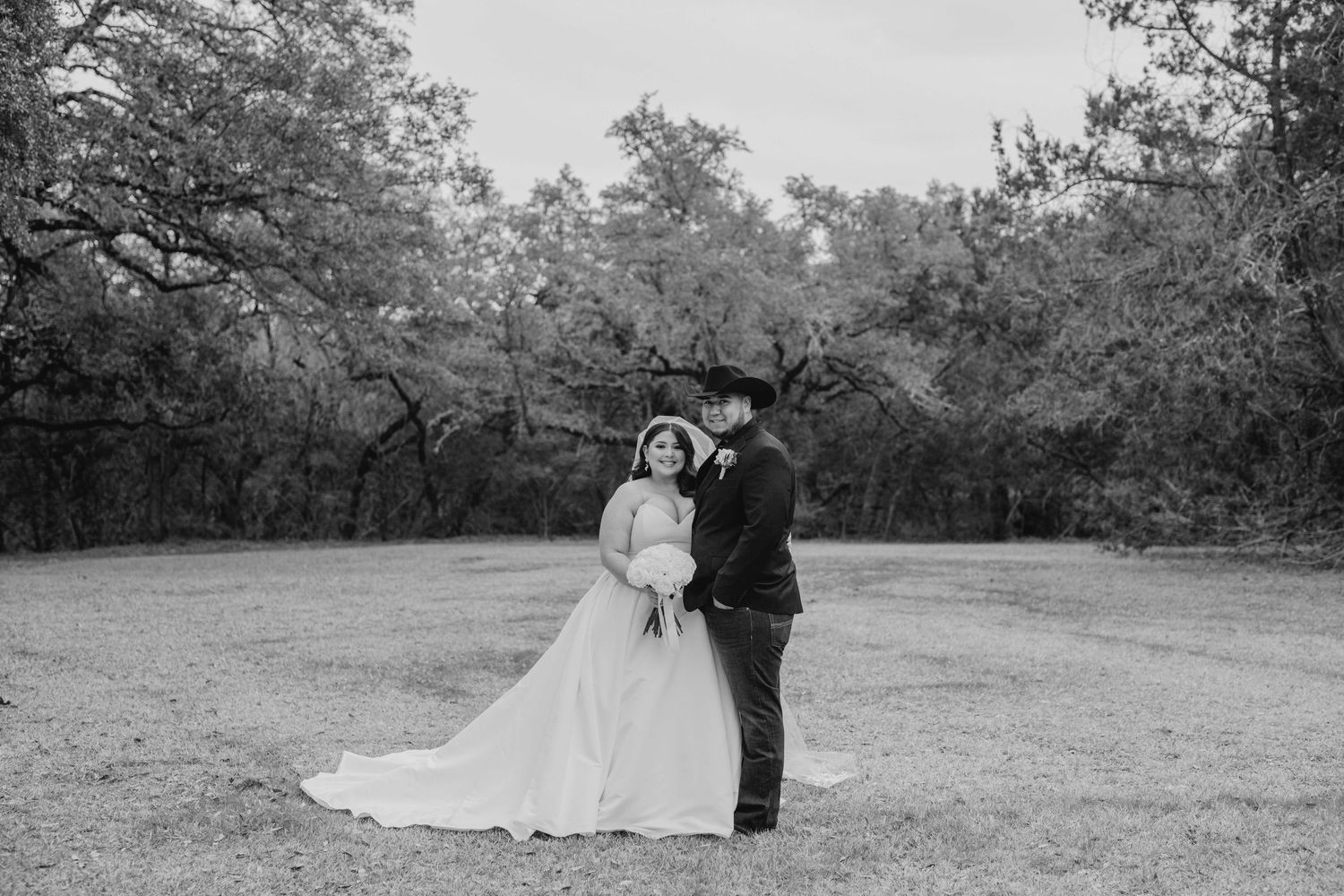 A couple in wedding attire embrace in a black and white photo at an outdoor venue surrounded by trees.