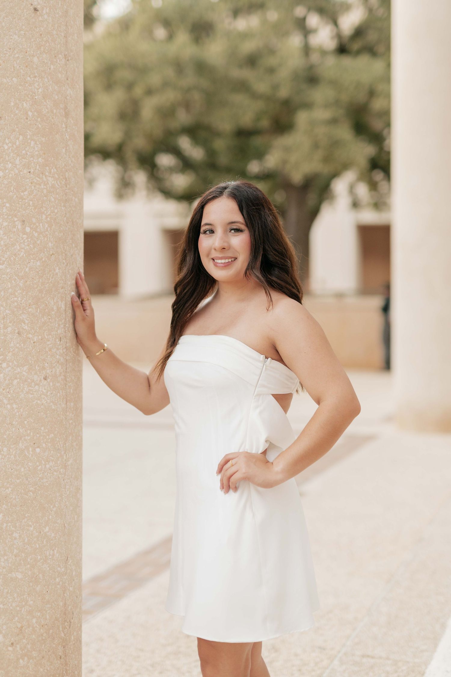 Graduate in white strapless dress poses by column during photo session.