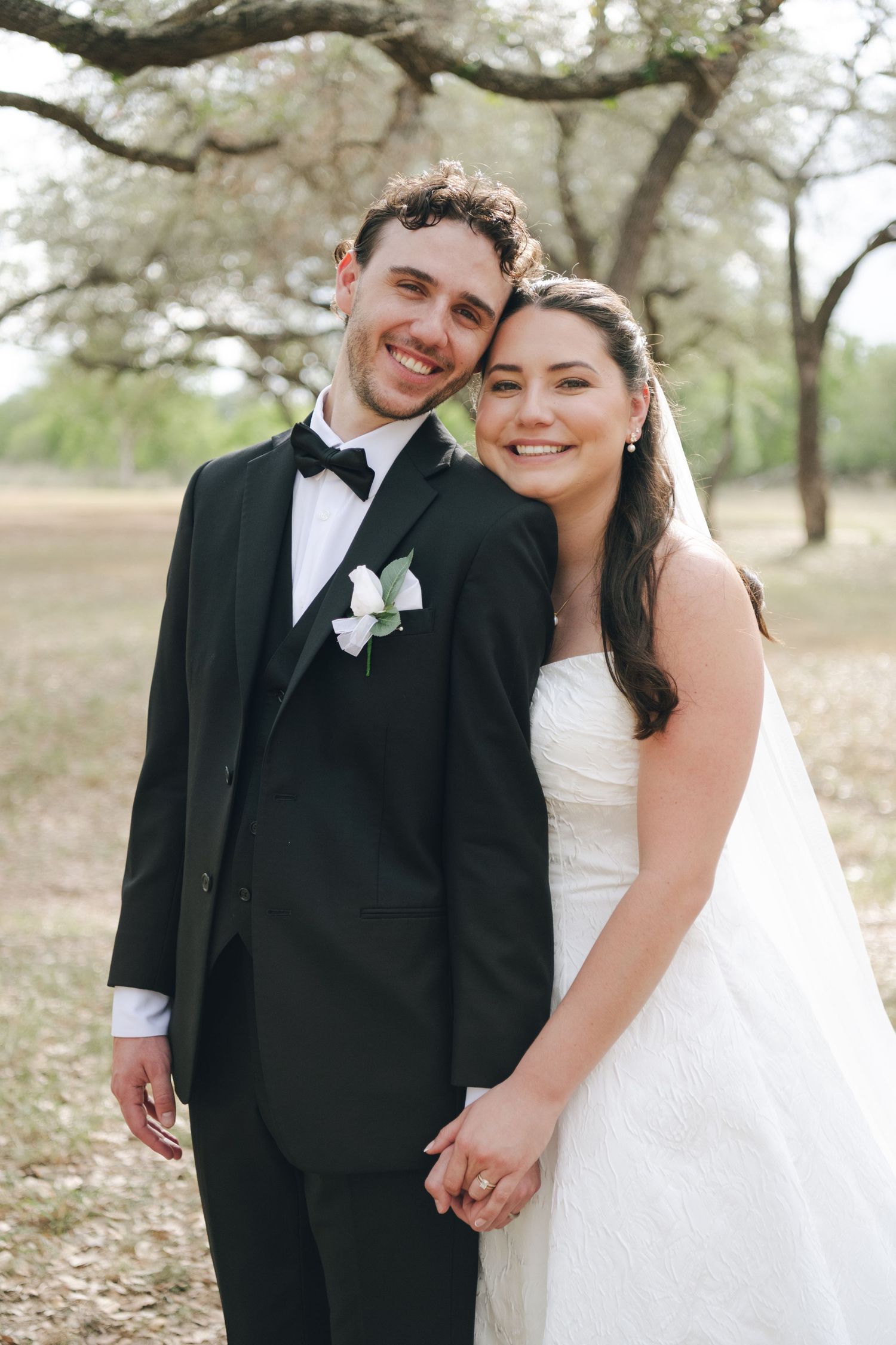 Smiling bride and groom pose outdoors under oak trees, she in a strapless white gown, he in a black tuxedo with boutonniere.