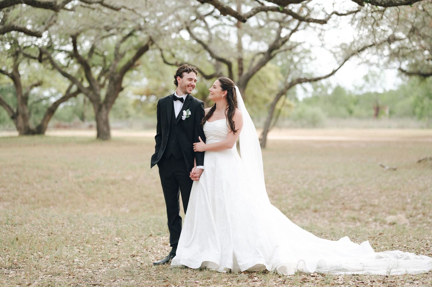 Bride and groom holding hands smiling at each other in a scenic outdoor field with large oak trees.