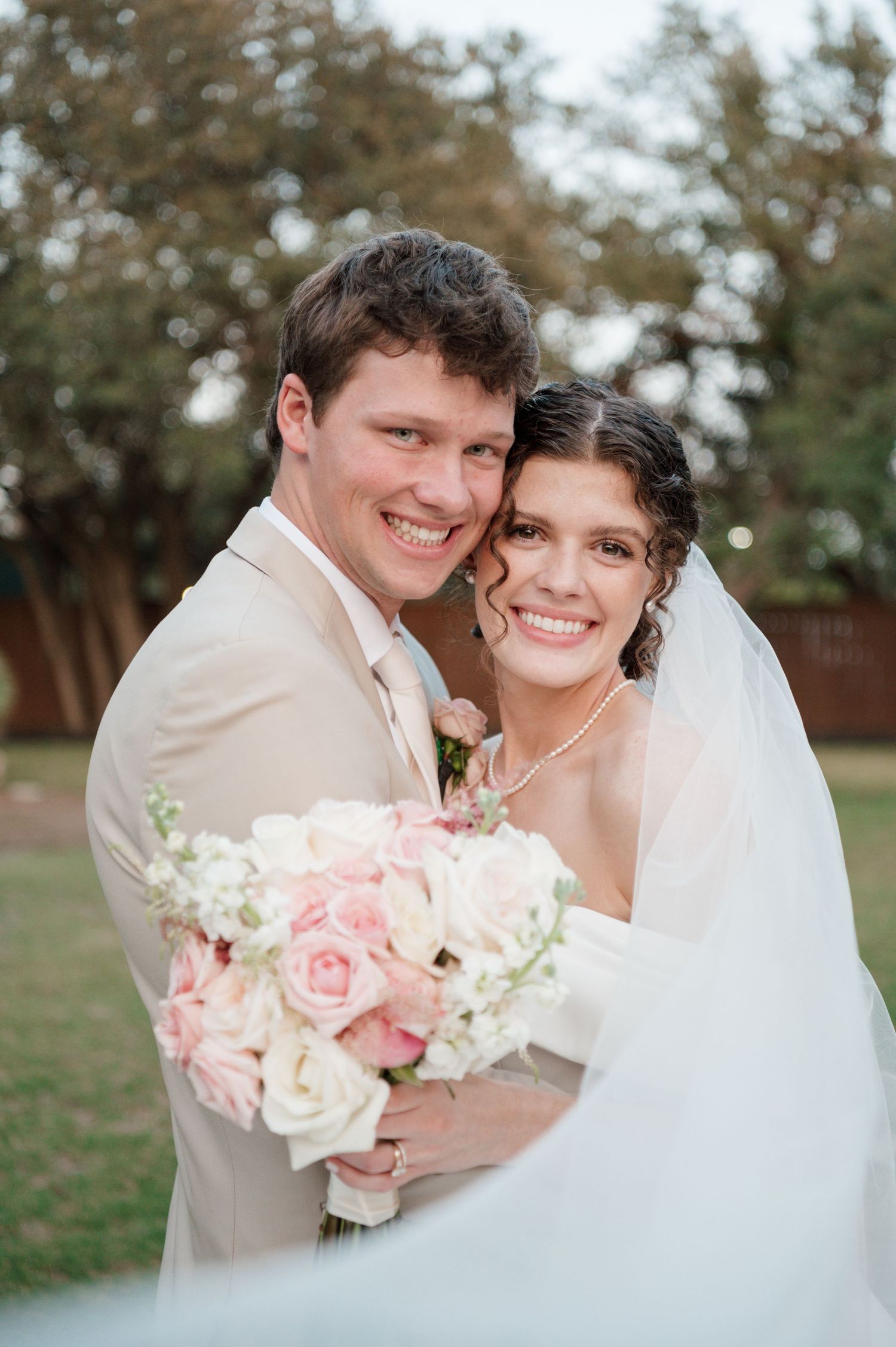 Newlywed couple embraces while holding pink and white rose bouquet outdoors.