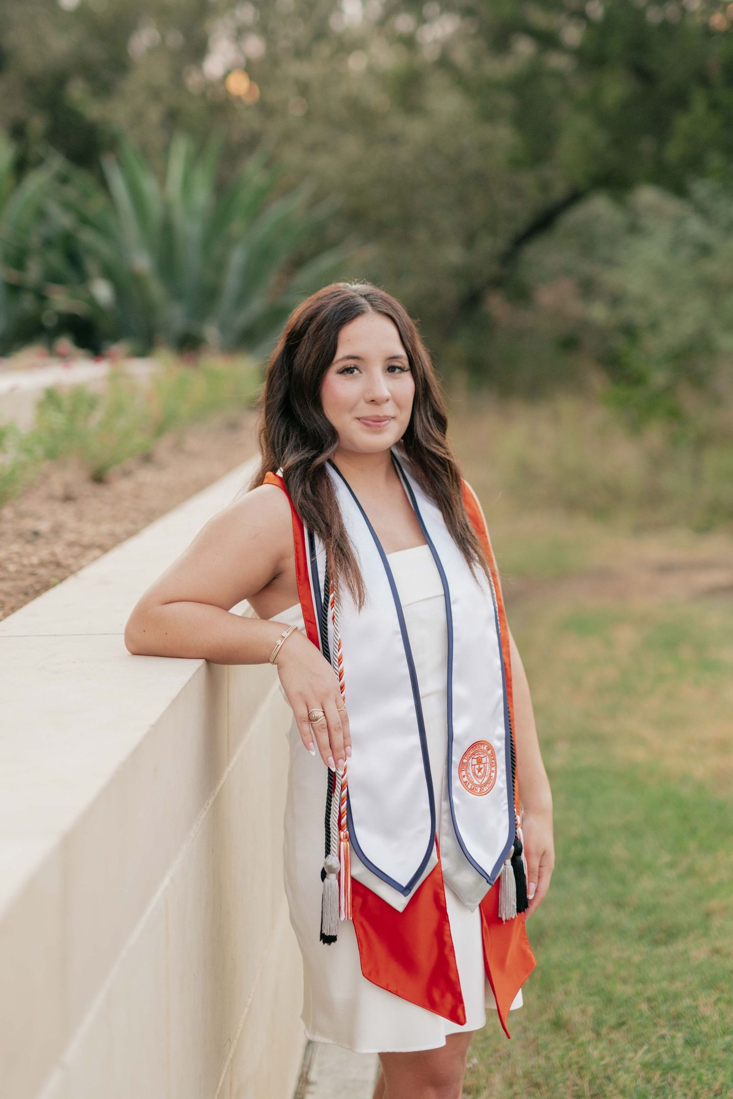 Graduate in white dress and decorative stole poses against retaining wall with desert landscaping.