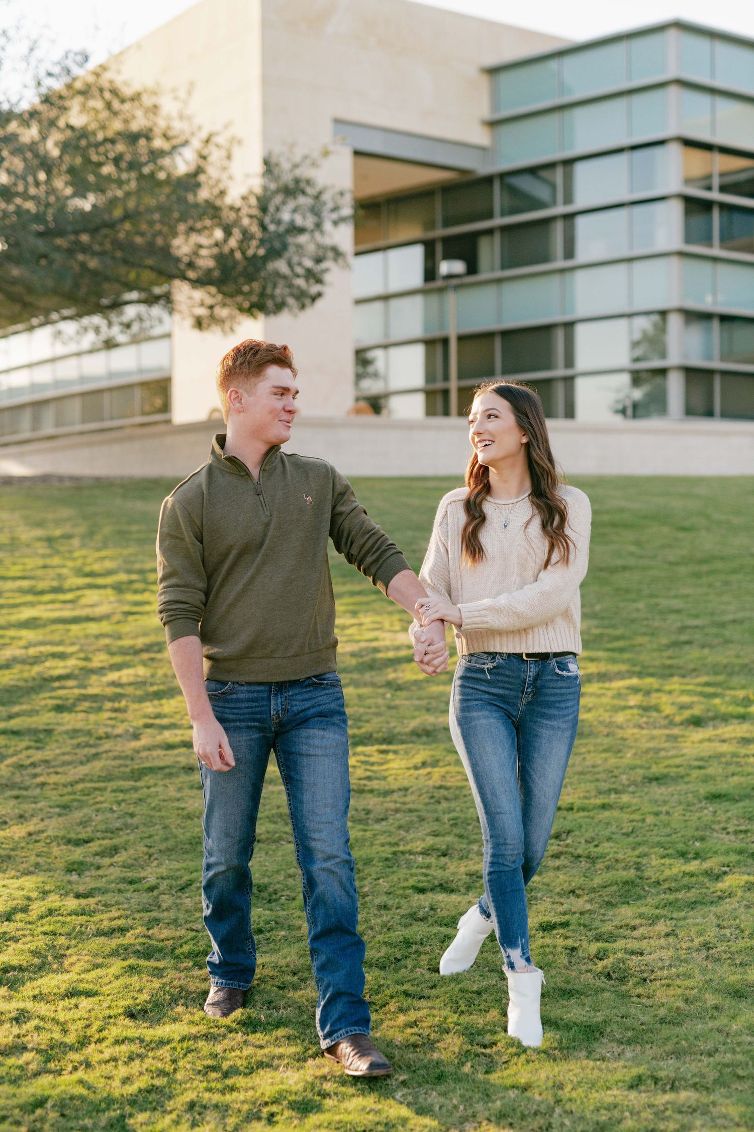 Couple walks together on a grassy slope with a modern building in the background.