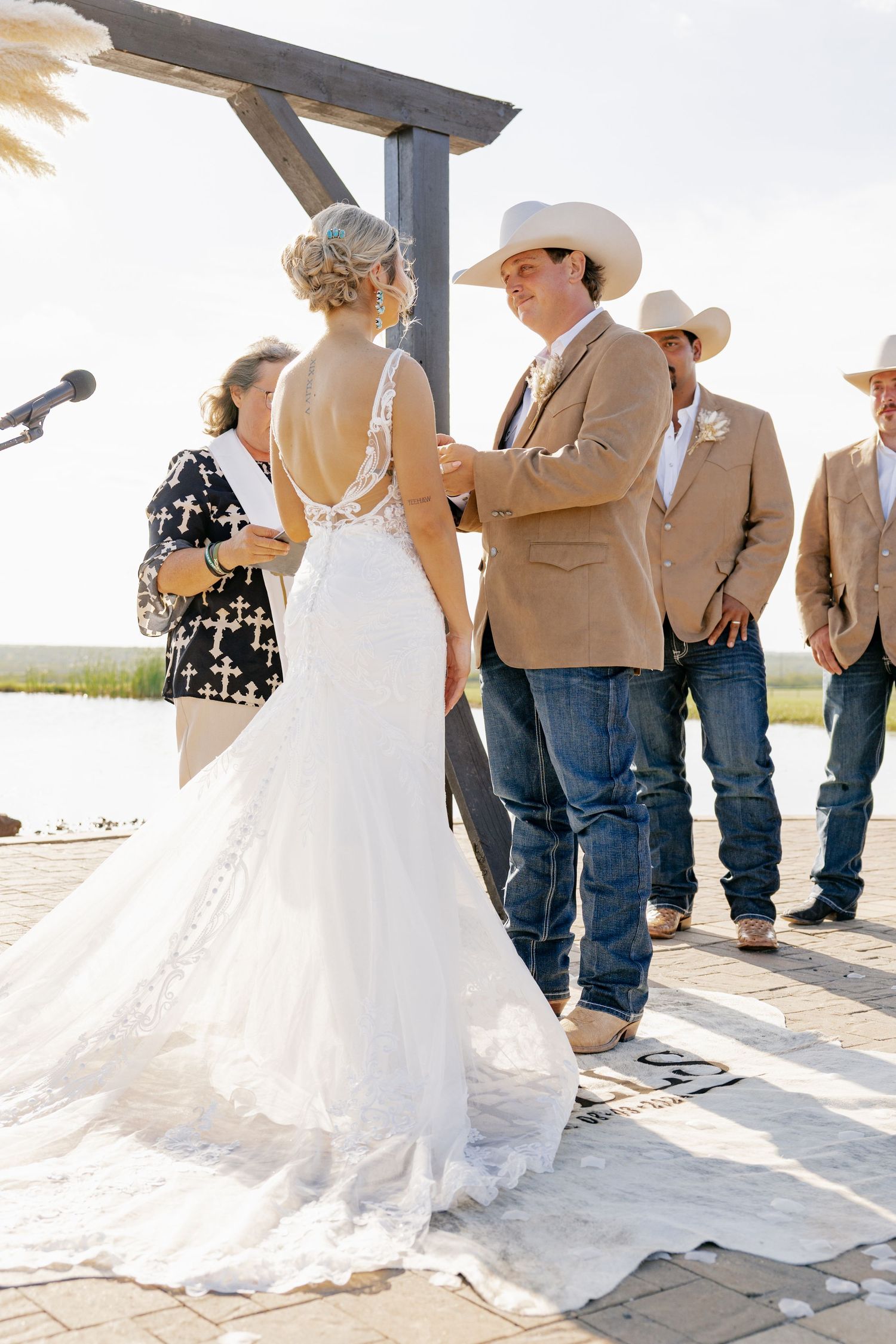 Outdoor wedding ceremony under wooden pergola with couple and wedding party in western attire.