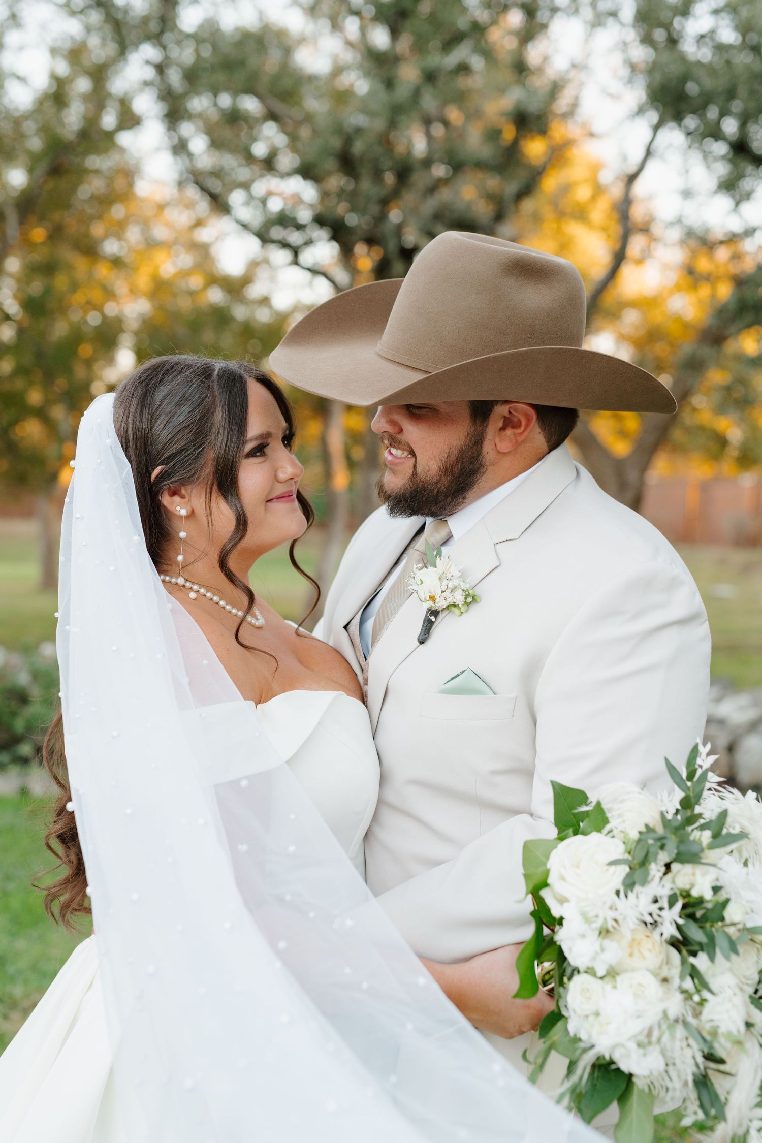 A couple in wedding attire share an intimate moment outdoors, with the bride holding white flowers and groom wearing a tan cowboy hat.