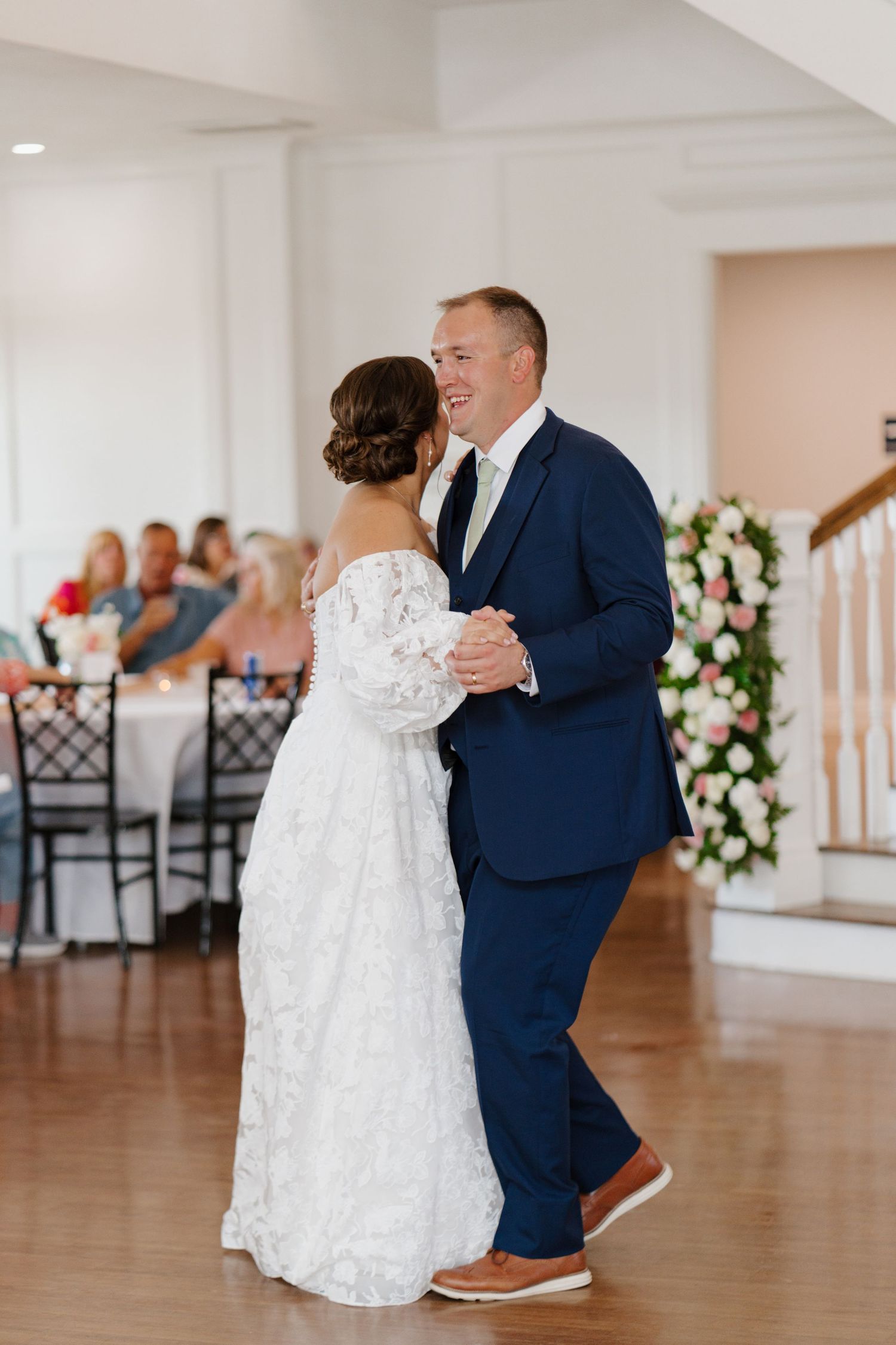 A couple shares a romantic dance together in a bright ballroom decorated with white floral arrangements.
