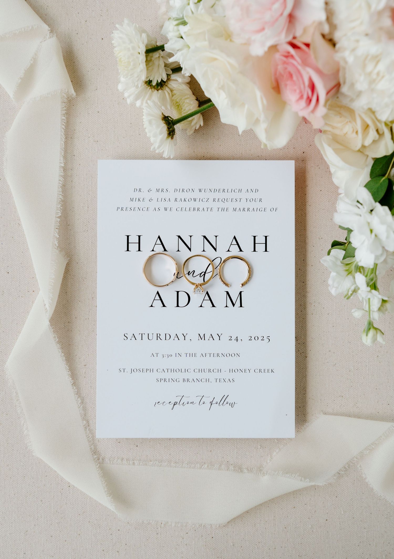 Wedding invitation and rings styled with delicate pink and white flowers on creamy textured fabric.