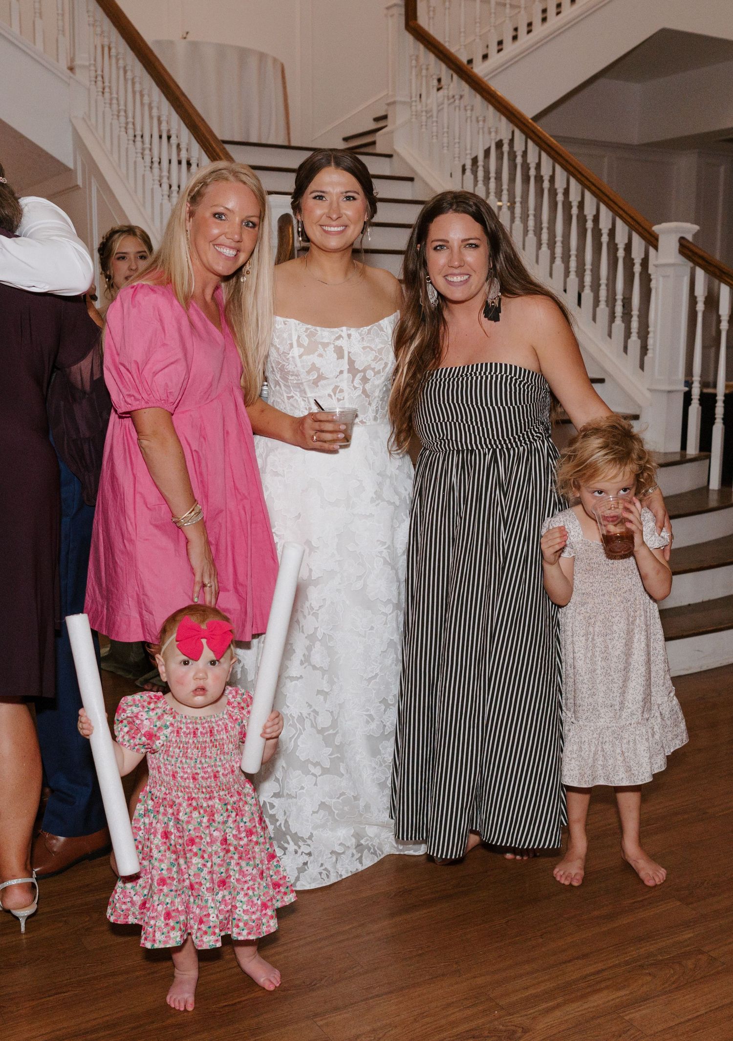Group of people in summer dresses standing together on hardwood floor near white staircase at indoor gathering.