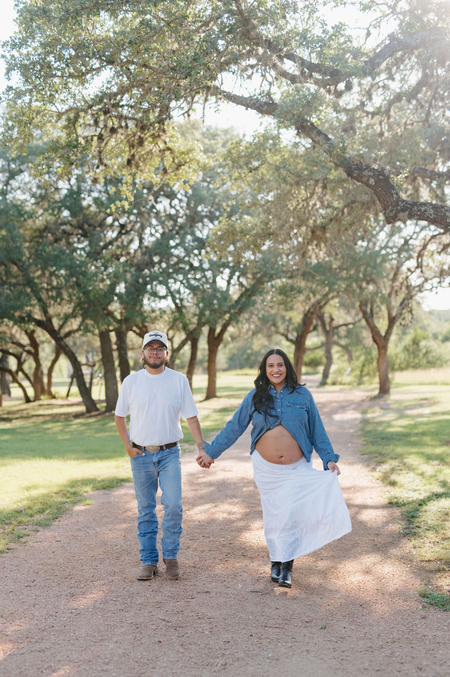 A couple walks hand in hand on a path through a sunlit oak tree grove during a joyful maternity photoshoot.