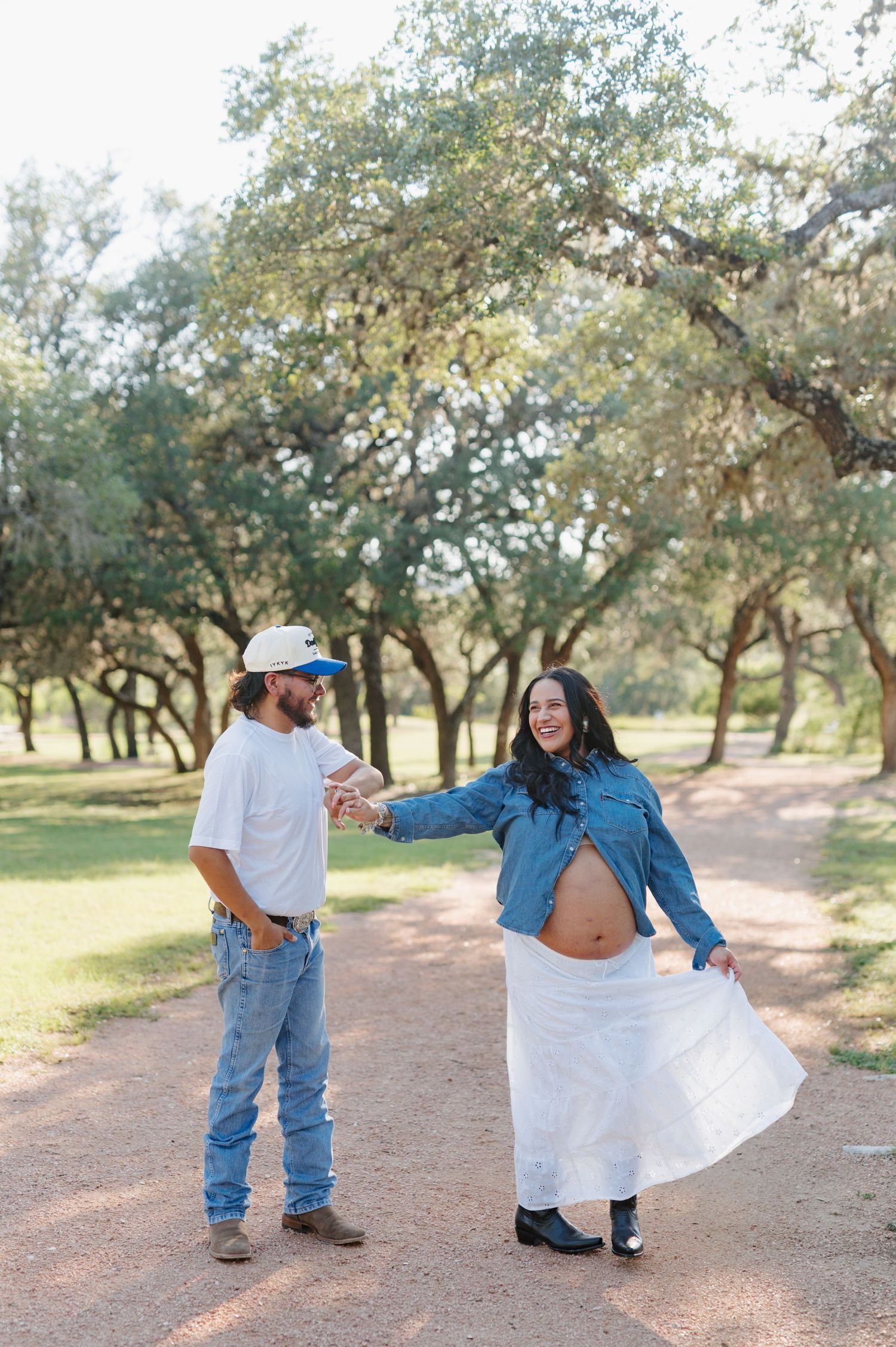 A couple in denim and white clothing poses playfully in a series of romantic photos under oak trees on a dirt path.