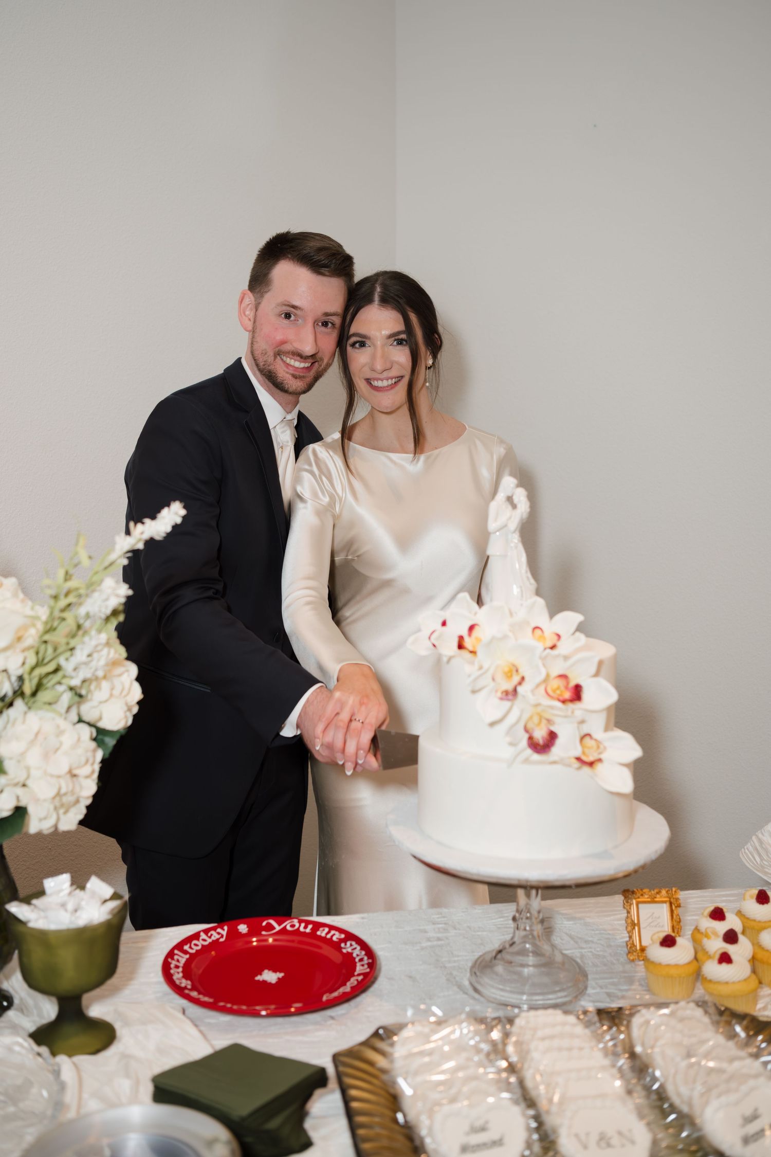 Newlyweds cutting their elegant white wedding cake surrounded by desserts and floral decorations.