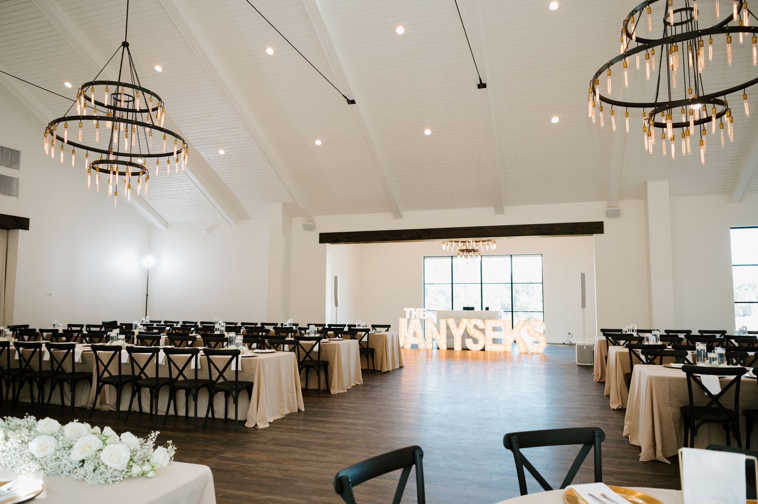 Modern white wedding venue interior with rustic chandeliers and cross-back chairs set up for an elegant reception.