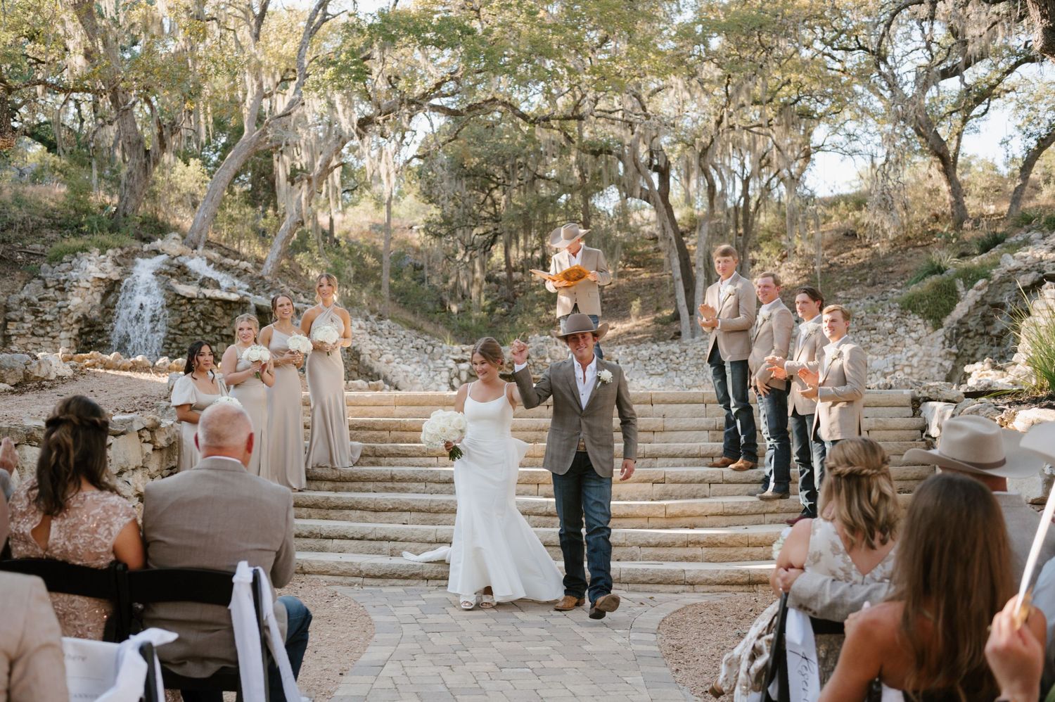 A wedding ceremony taking place on stone steps surrounded by trees and guests.