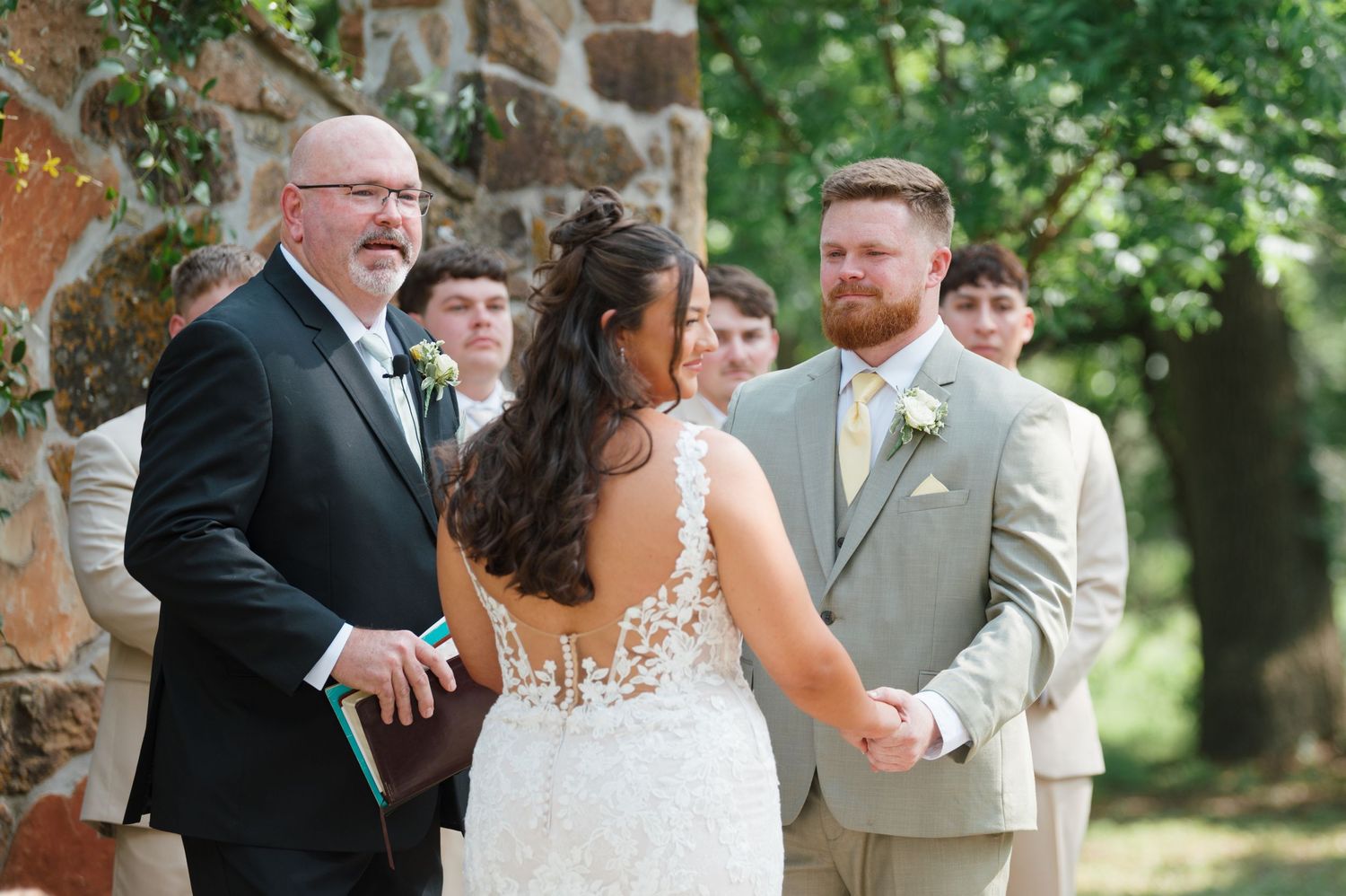 Outdoor wedding ceremony with bride in lace gown holding hands with groom in tan suit, officiant smiling nearby.
