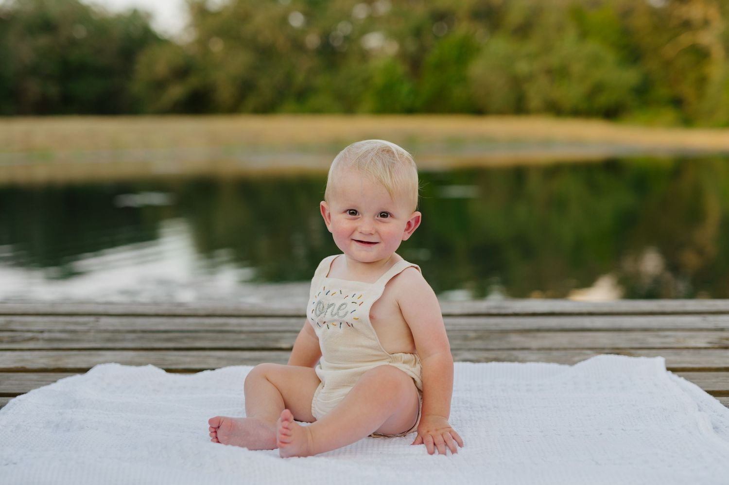 A baby in a white outfit sits on a dock by a peaceful lake during sunset.