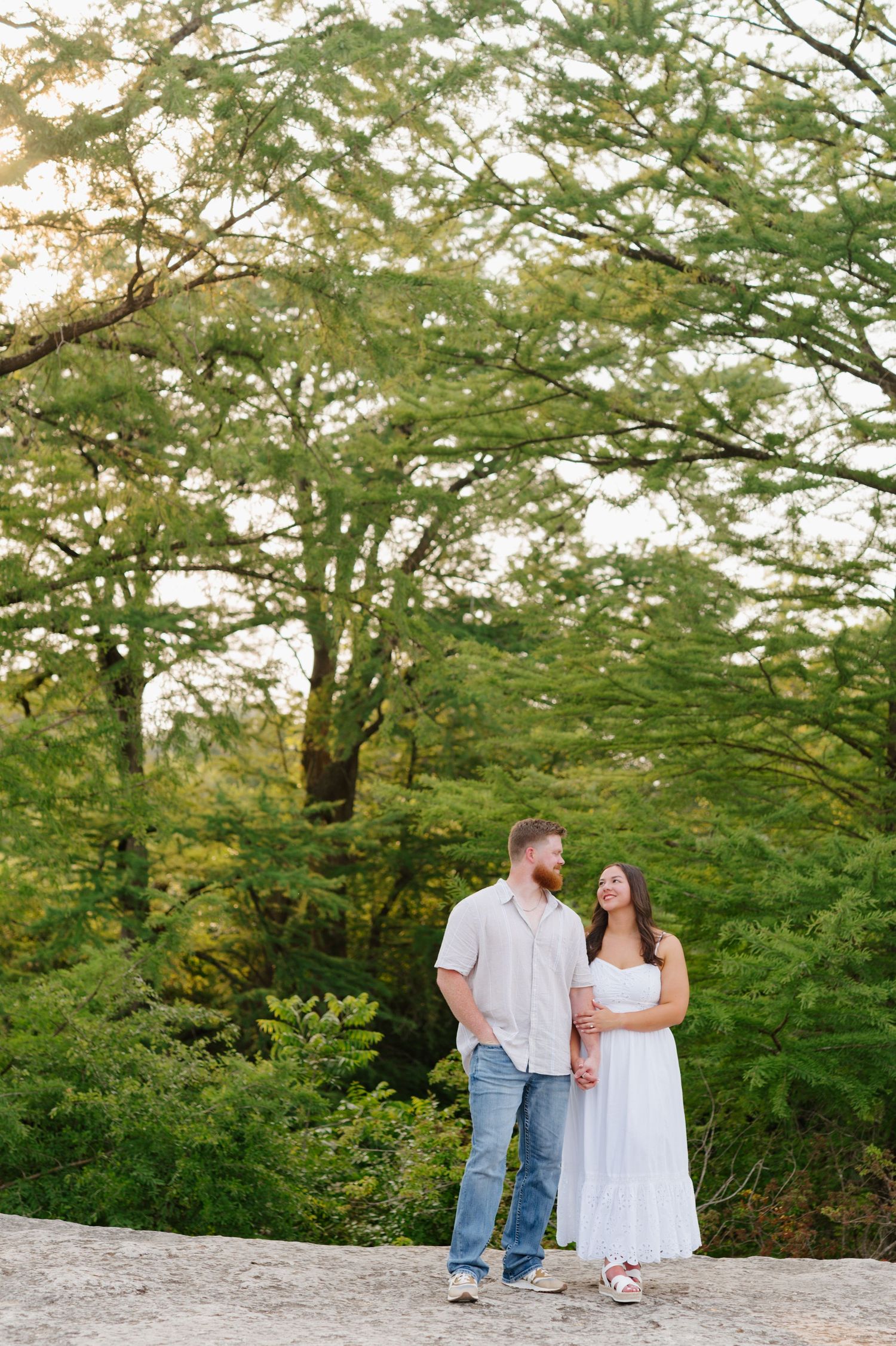 A couple shares a romantic moment while walking together on a path surrounded by lush green trees on a sunny day.