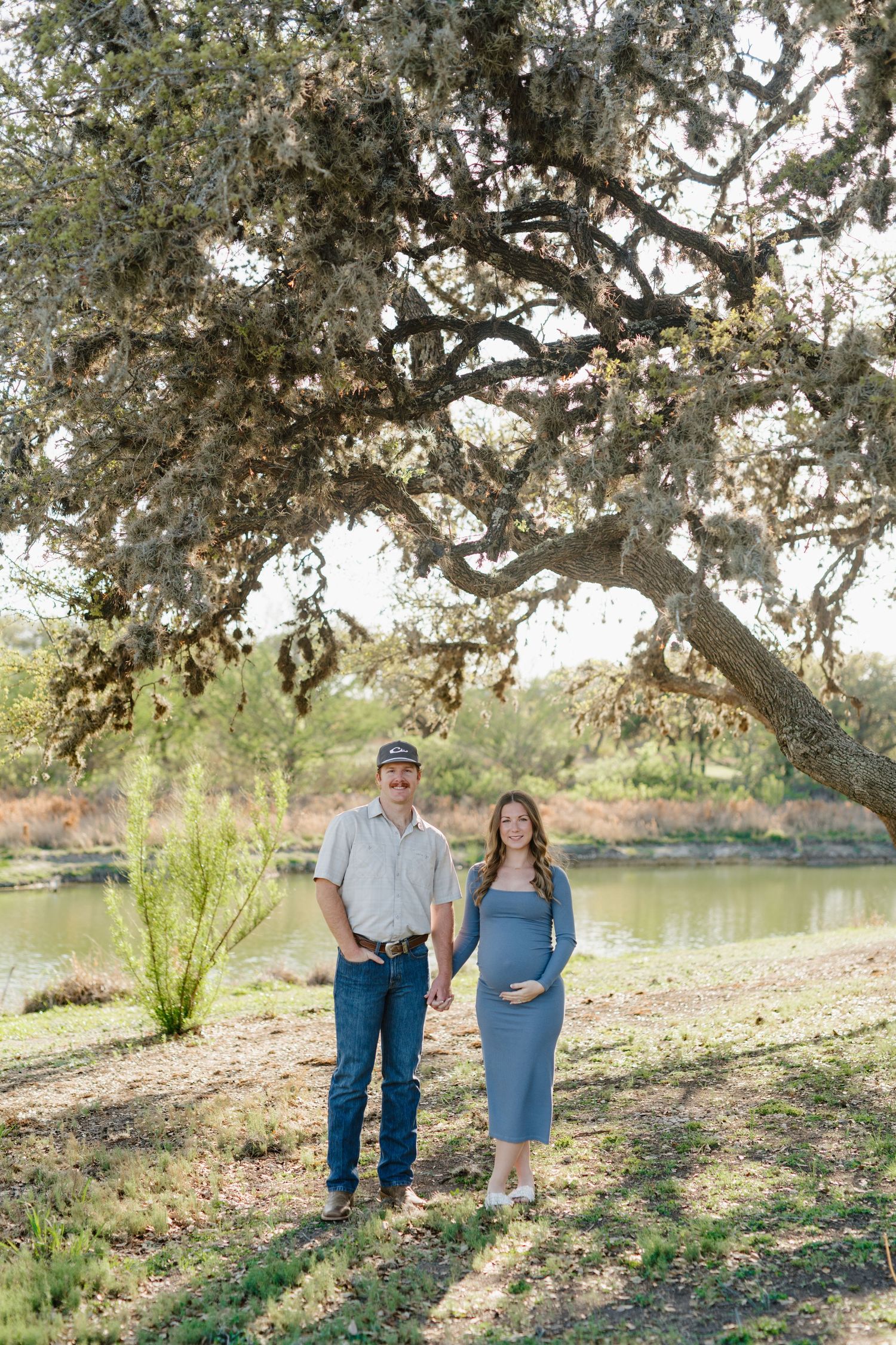 An expectant couple poses beneath a sprawling oak tree near a peaceful lake.