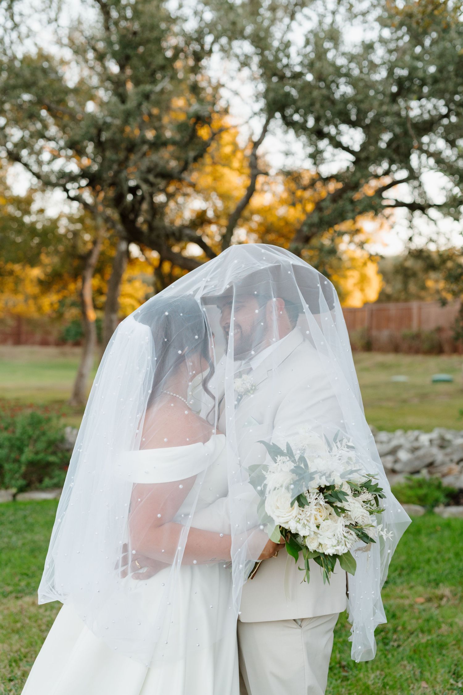 A white floral bridal bouquet held beneath a flowing veil against a backdrop of autumn trees in a garden setting.